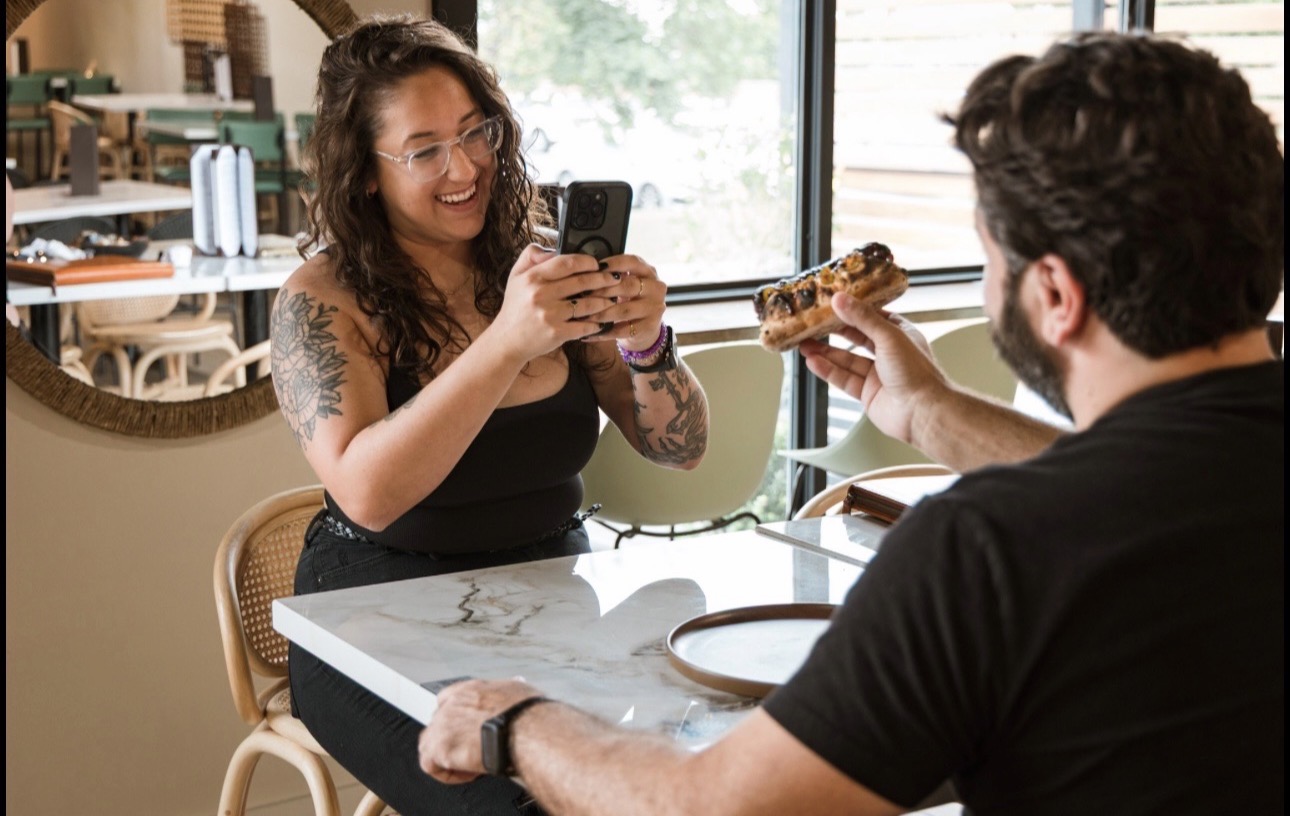 Woman taking a photo of a man holding a sandwich at a table in a cafe, smiling, with a window in the background.