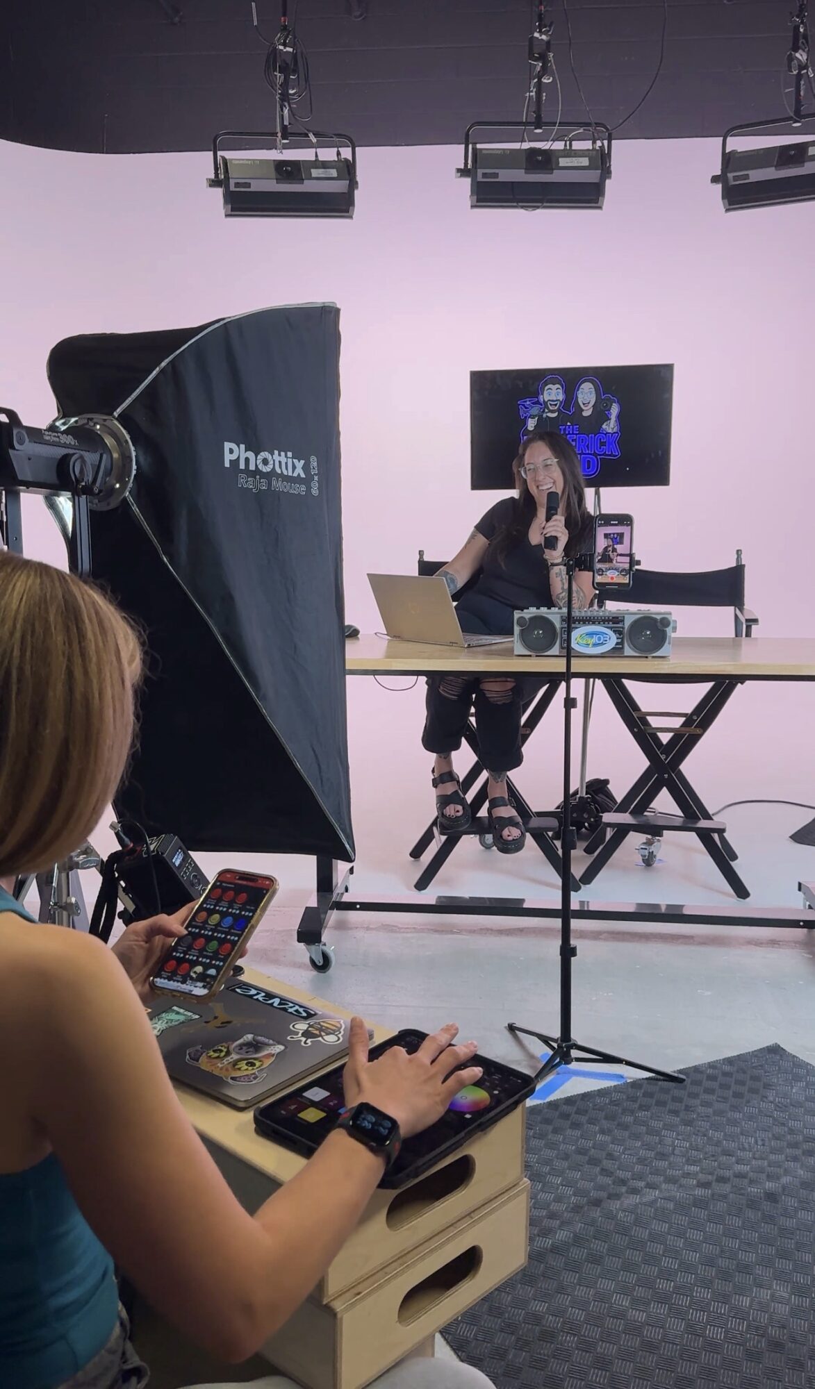 A woman performs on a stage with a DJ setup, a screen, and studio lighting, while a person operates a camera in the foreground.