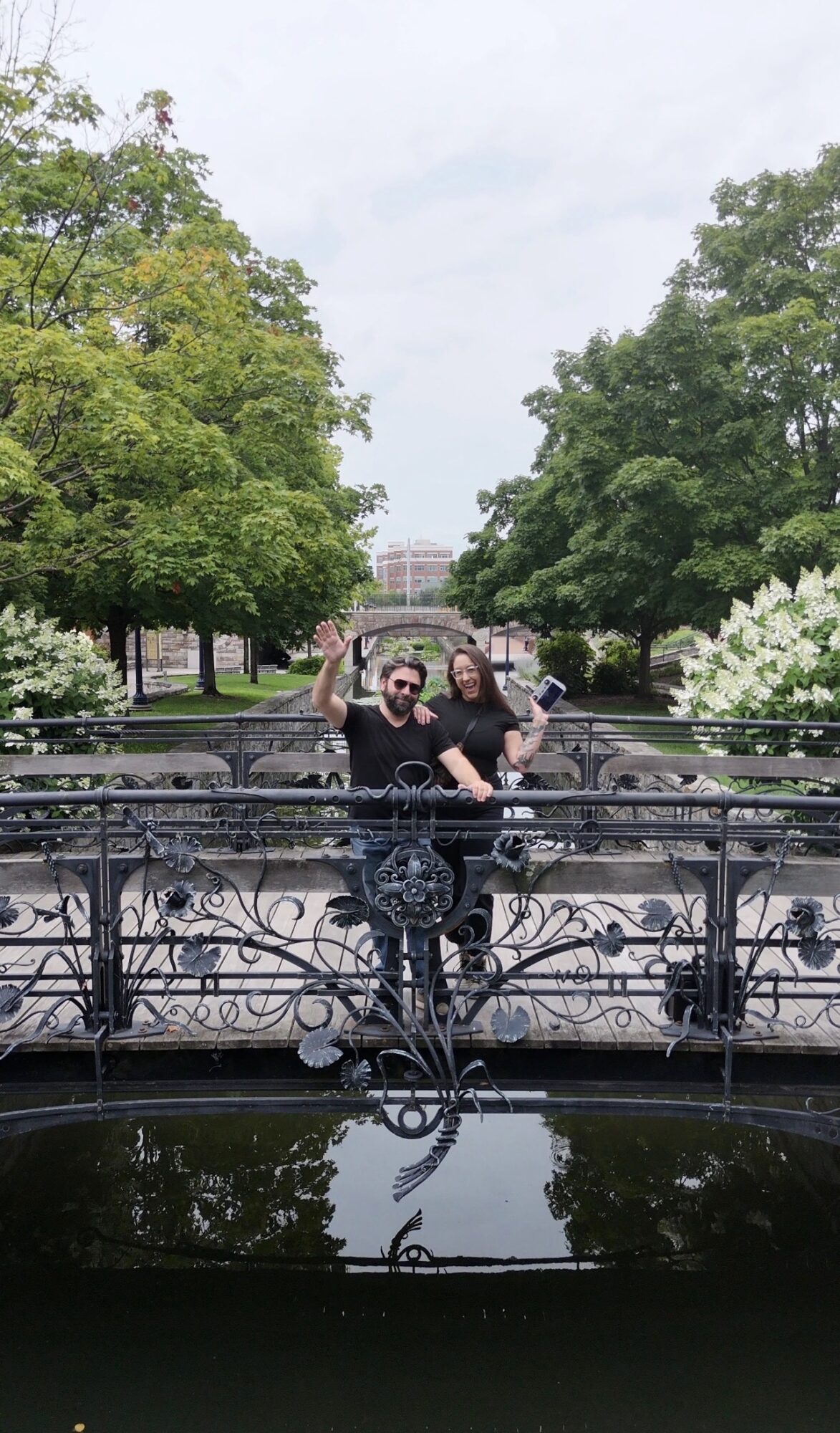 Two people stand on a decorative bridge with trees and water in the background, one making a peace sign, the other smiling.