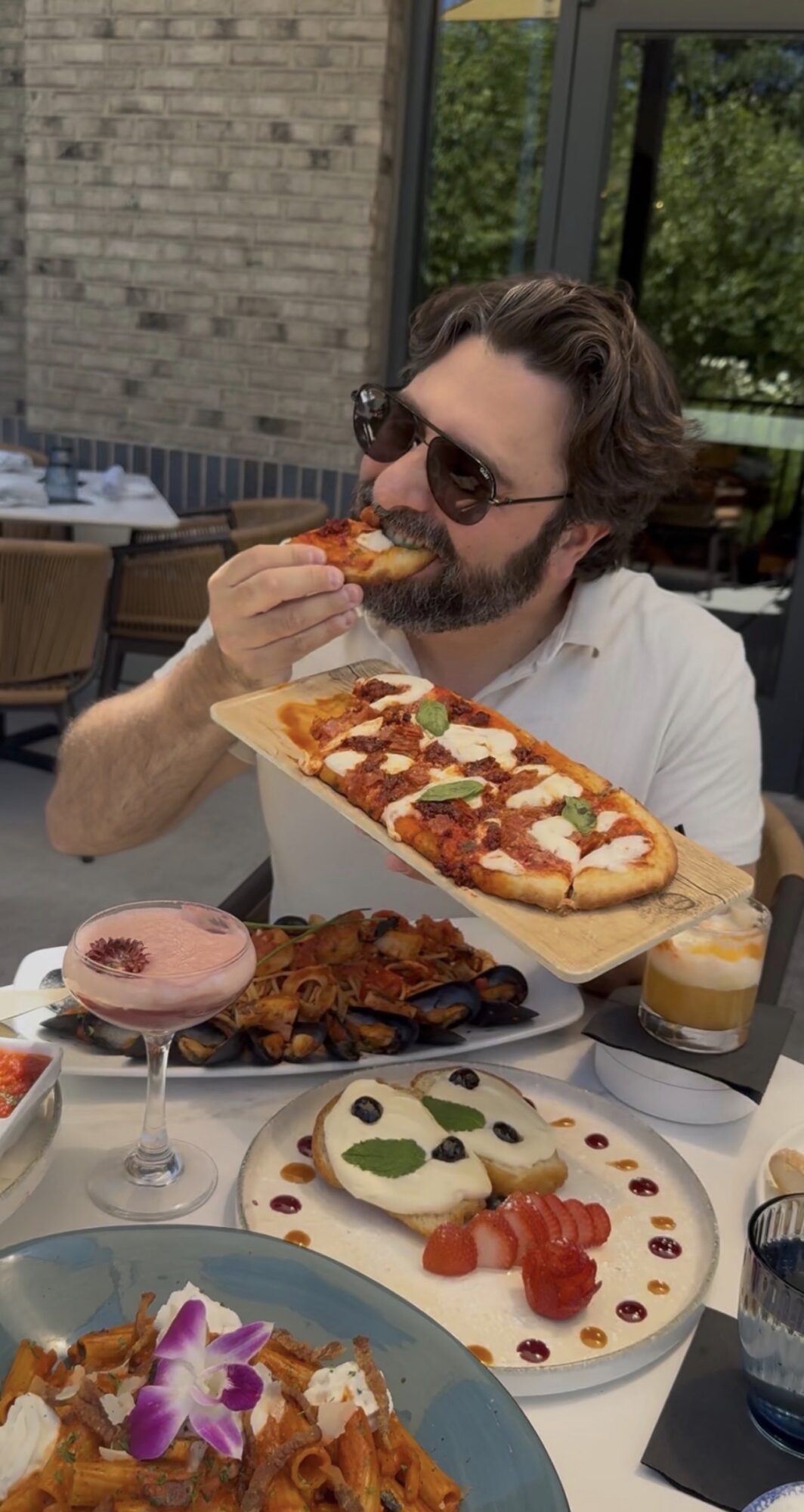 Man with sunglasses eating pizza at outdoor table with various desserts and food, brick wall and greenery in background.