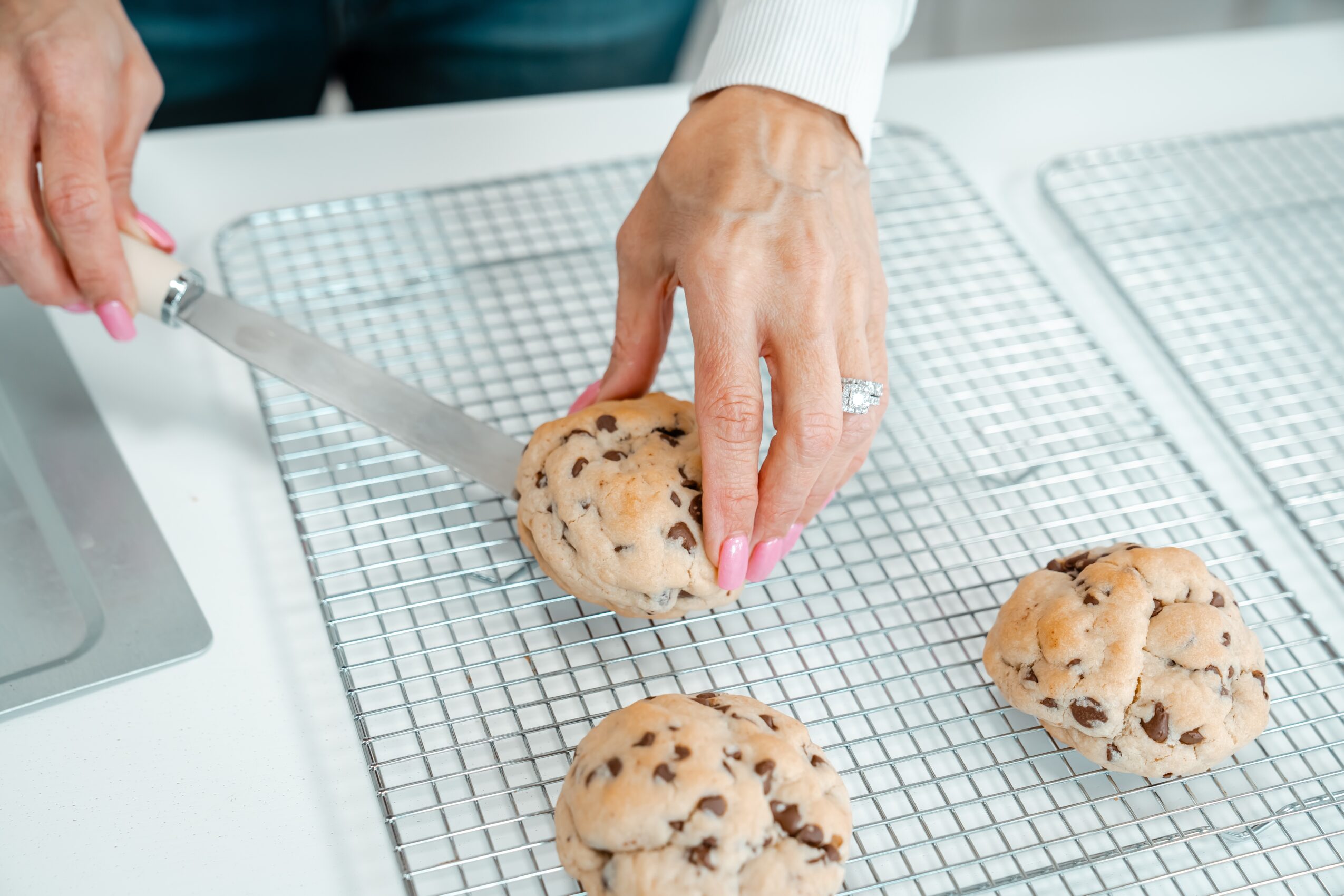 Person cutting a chocolate chip cookie on a wire rack with a knife, three cookies on the rack, white background