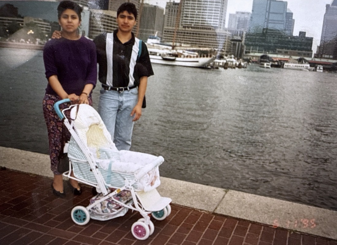 Two women stand by a stroller near a river with city buildings in the background.