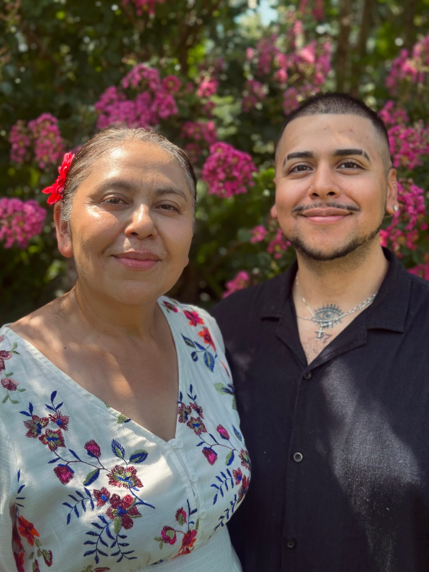 Two people, a woman and a man, stand outdoors in front of pink flowering bushes, smiling at the camera.