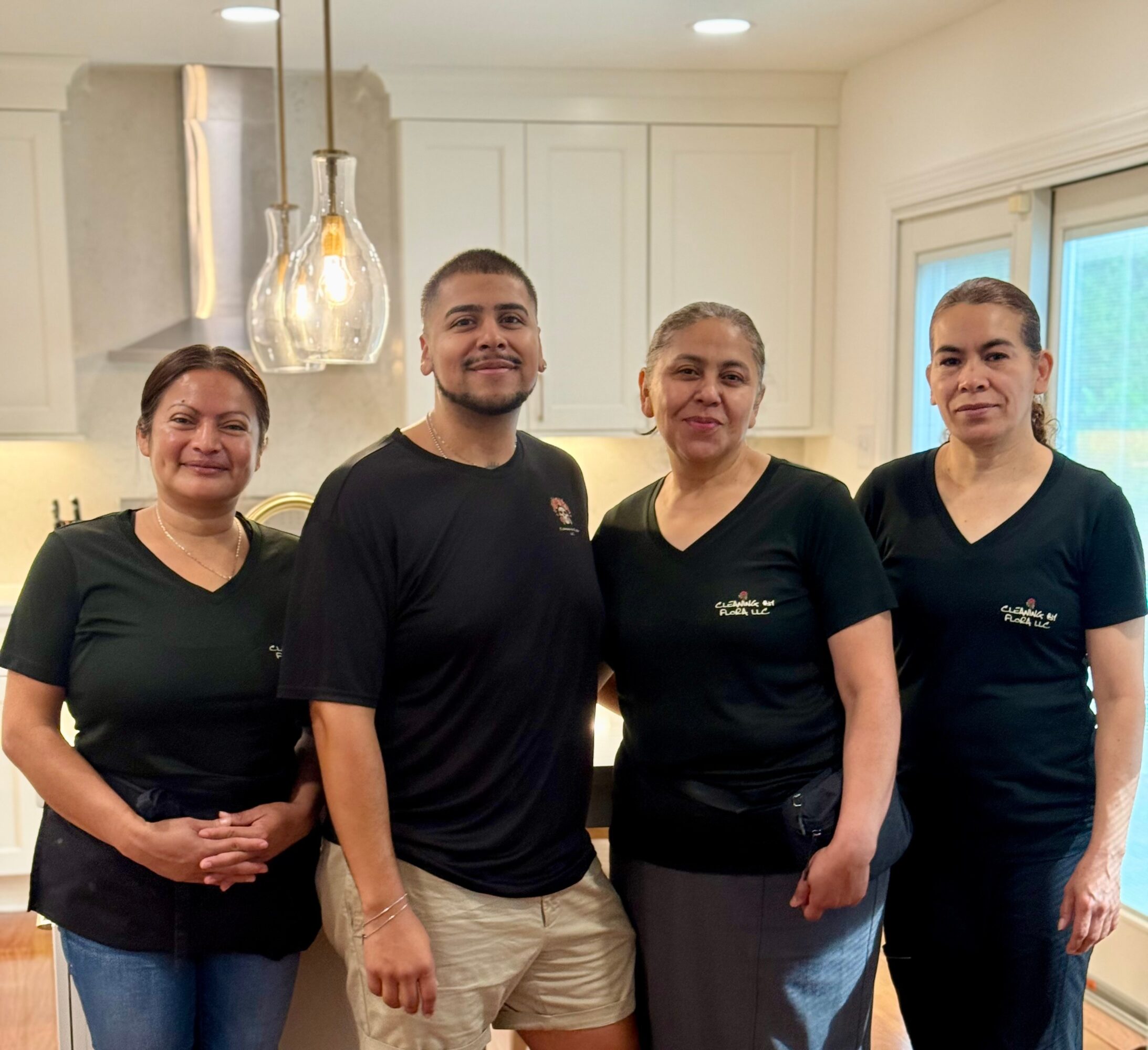 Four people standing in a kitchen, smiling, with pendant lights and a window in the background.