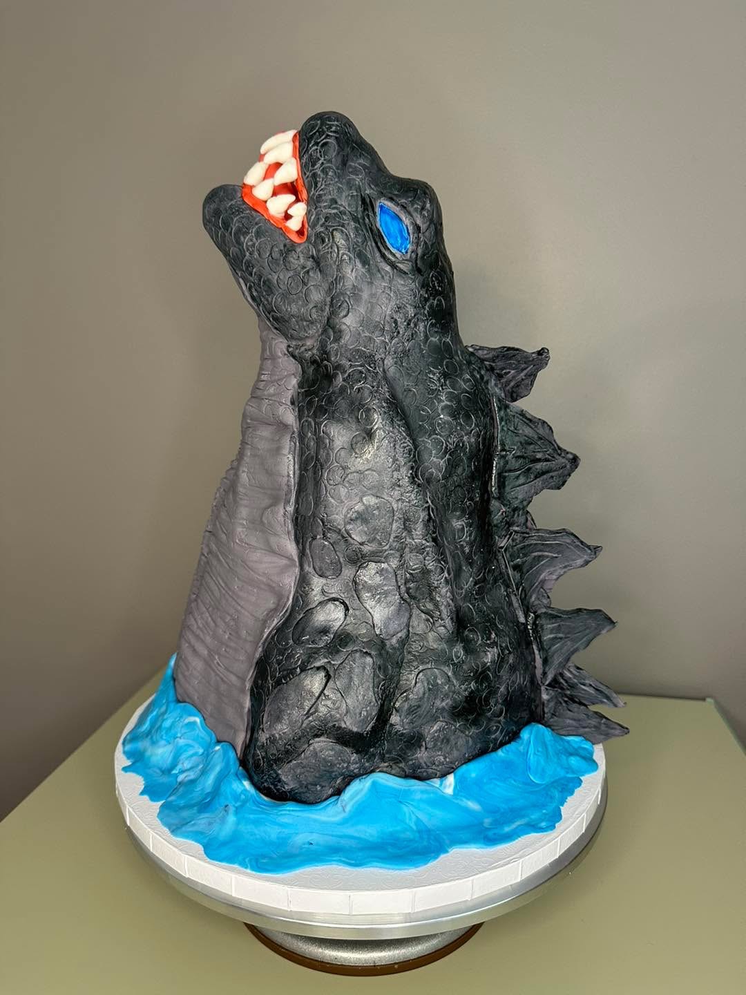 Cake shaped like a dinosaur with an open mouth, blue eyes, and textured black body, on a white base with blue icing.