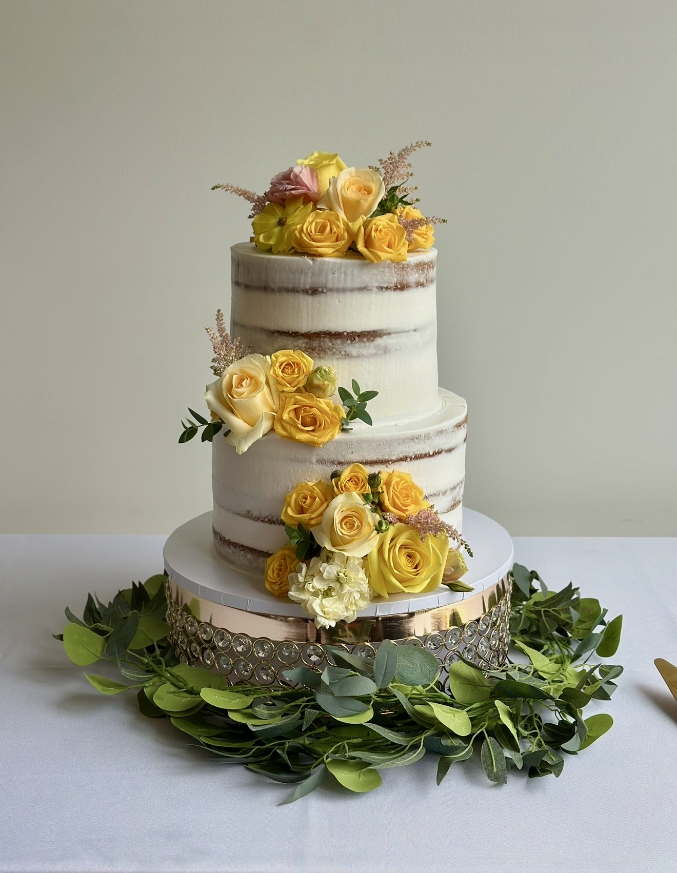 Two-tiered naked cake decorated with yellow roses and greenery, on a white table with a green leafy base.