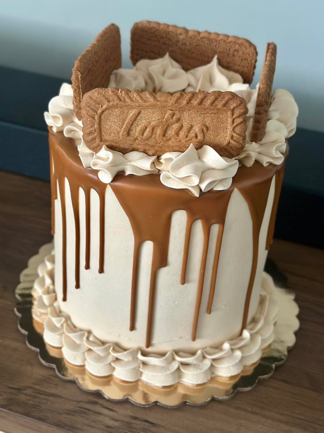 Cake with chocolate drip, whipped cream, cookies, and a Lotus biscuit on top, on a gold base.