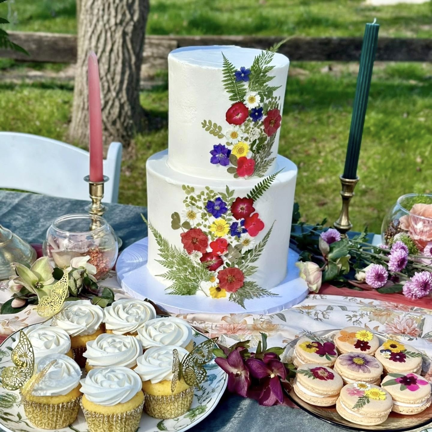 Two-tiered white cake decorated with colorful flowers and greenery, surrounded by cupcakes, flowers, and candles outdoors.