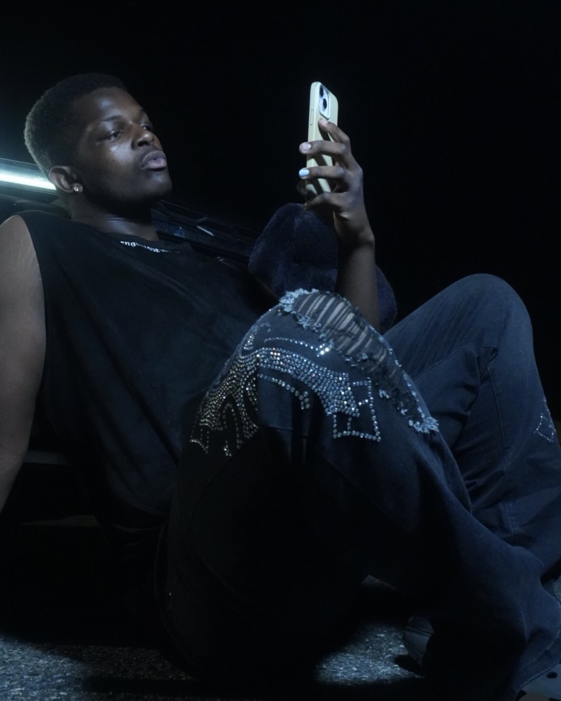 Person sitting in dark environment, looking at phone, wearing dark clothing and jeans, with a light source behind them.