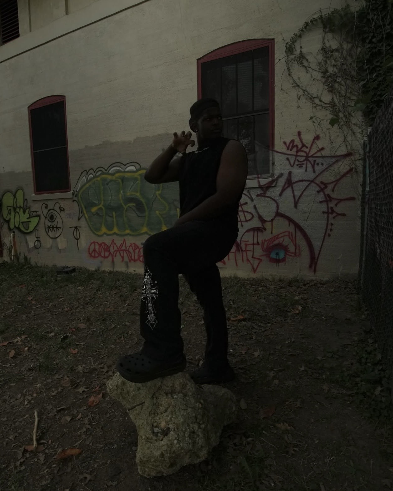 Person sitting on a rock outdoors, raising hand, with graffiti-covered wall behind them, in low light.