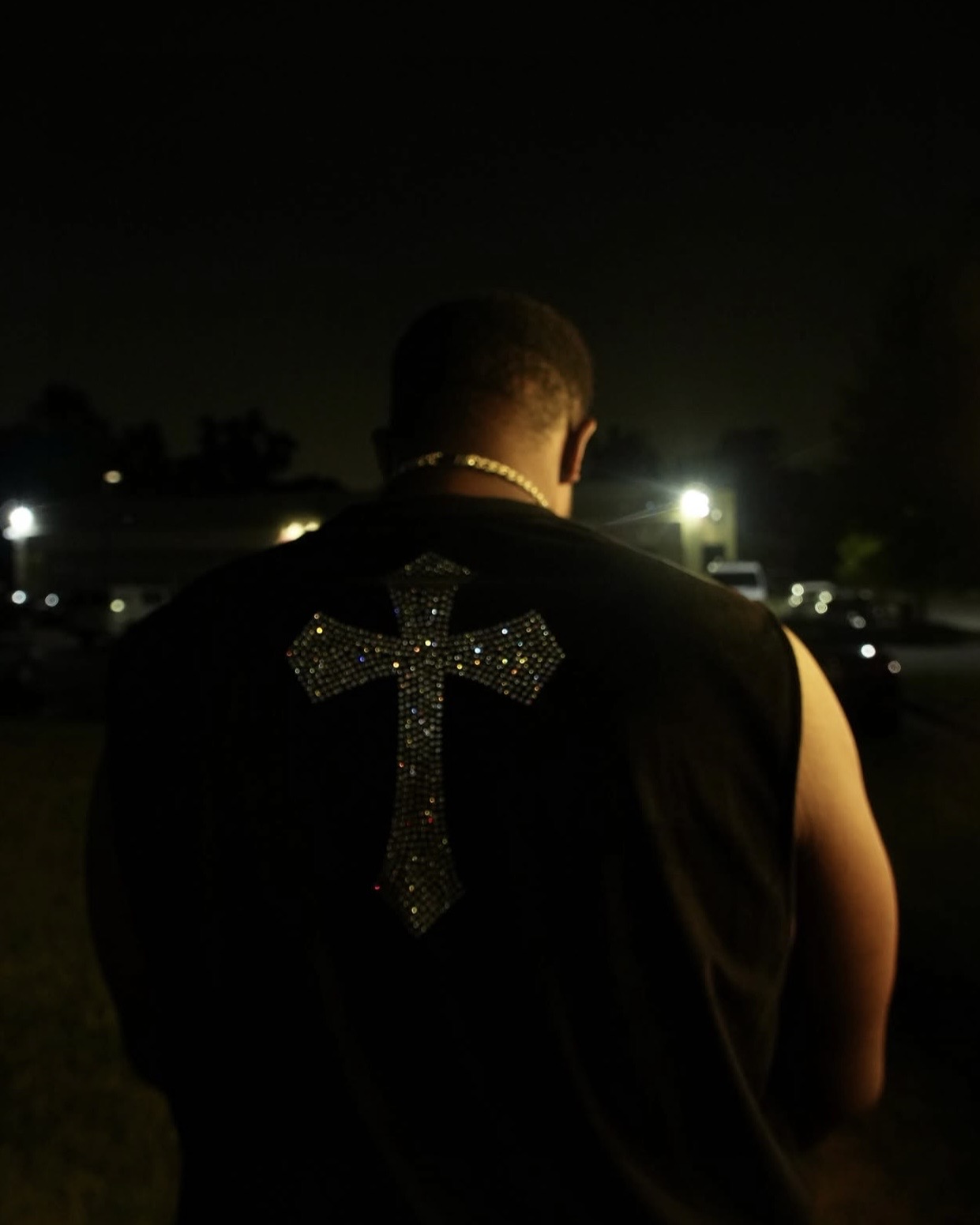 Person with back facing camera, wearing a sleeveless shirt with a glittery cross on the back, nighttime setting