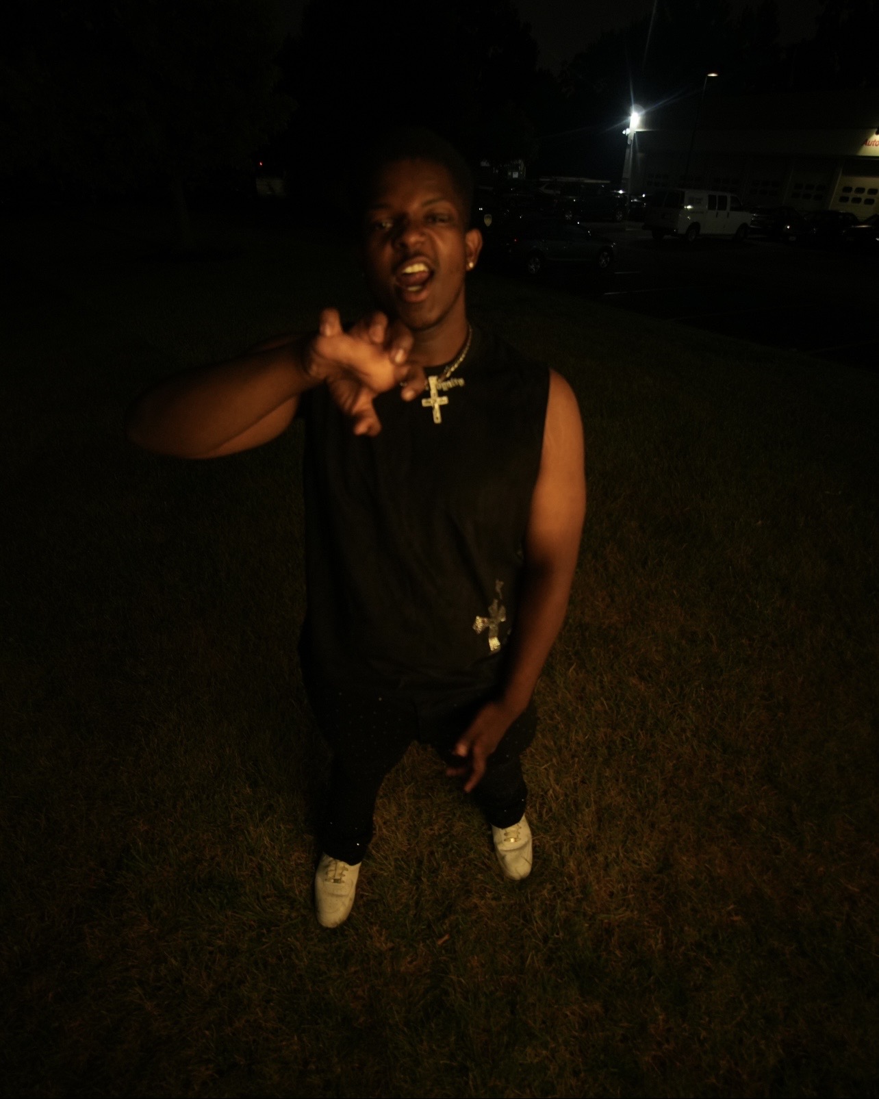 Person standing outdoors at night, gesturing with hand, wearing a sleeveless top, necklace, and sneakers, with dark background.