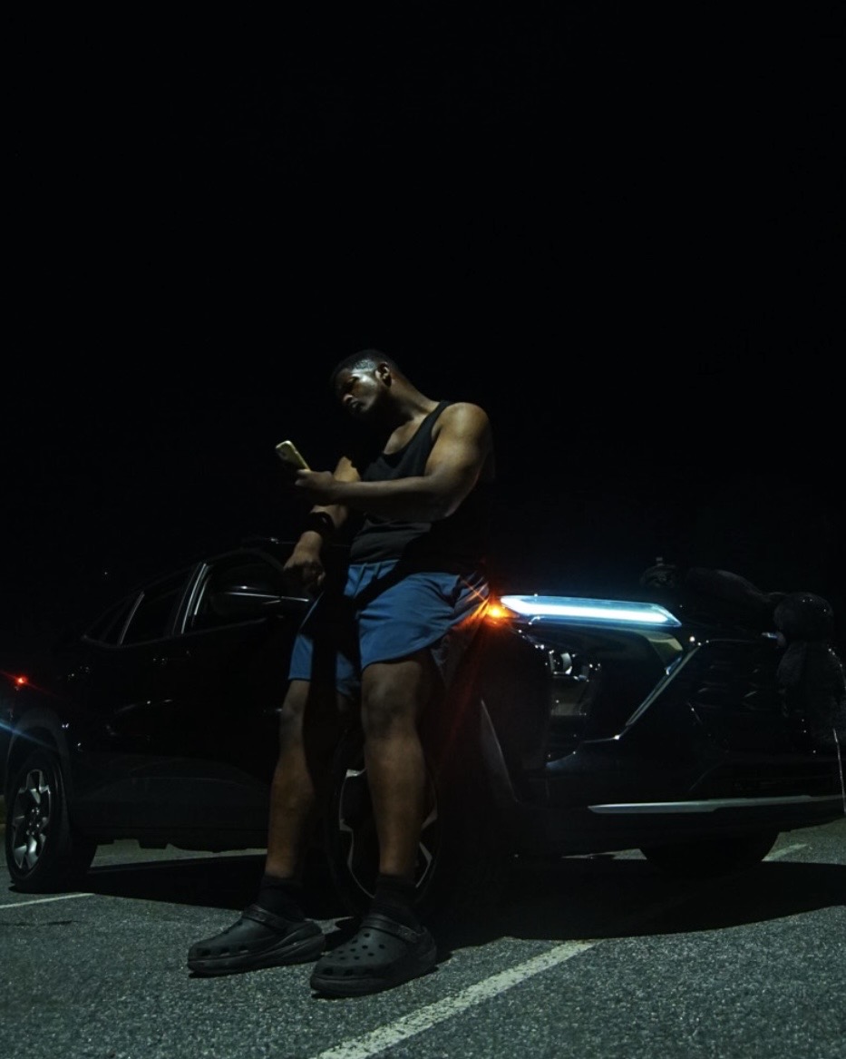 Young man standing next to a car at night, looking at his phone, wearing sunglasses, tank top, and shorts.