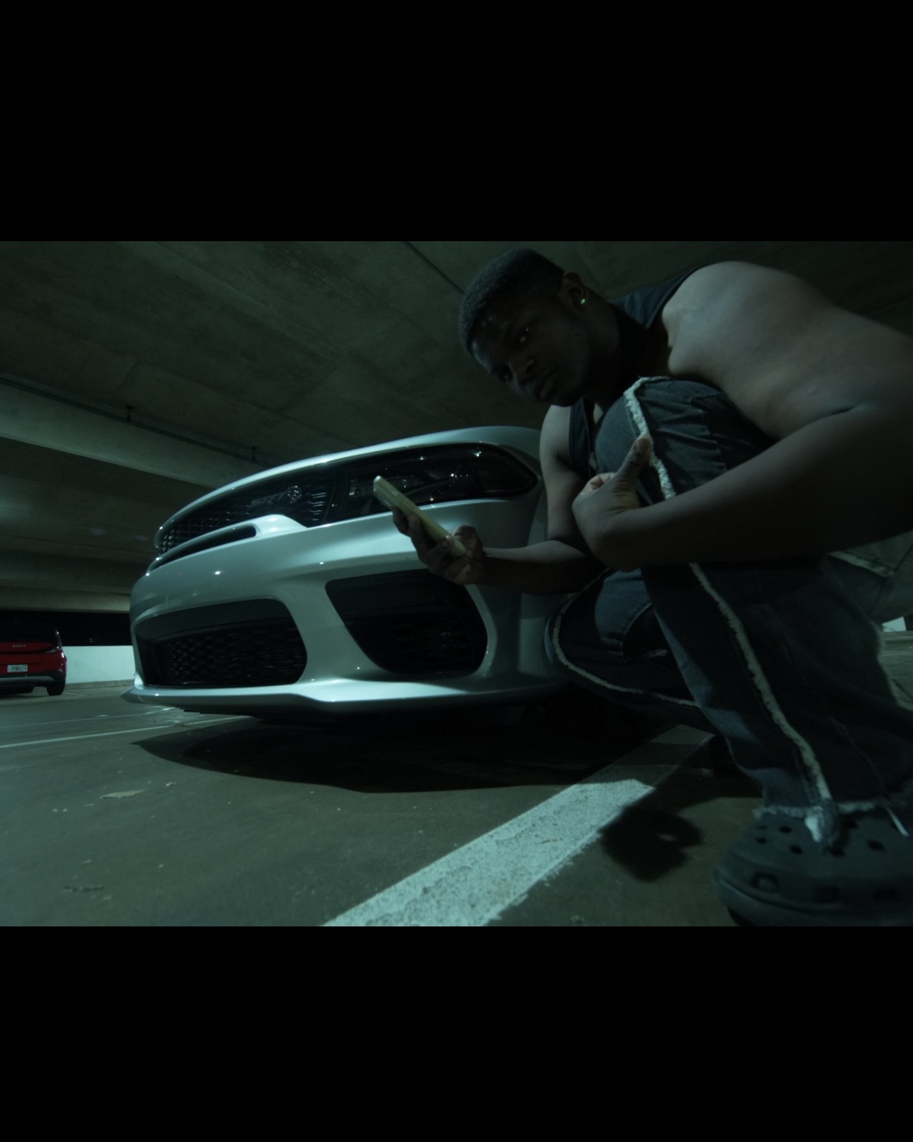 Person crouching next to a white sports car in a parking garage, looking at a phone, with a dark ceiling overhead.