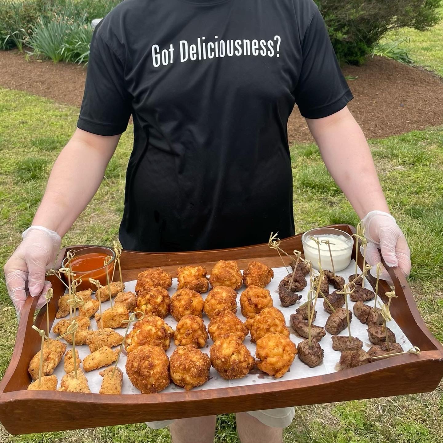Person holding a tray of fried chicken and skewered meat outdoors, wearing a black shirt with white text, in a garden setting.