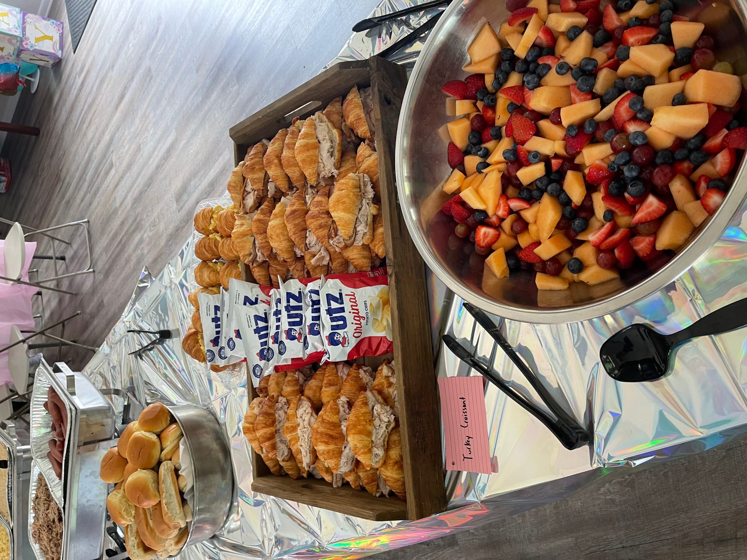 Table with assorted pastries, a bowl of fruit salad, and a carton of milk, with a pink note card nearby.