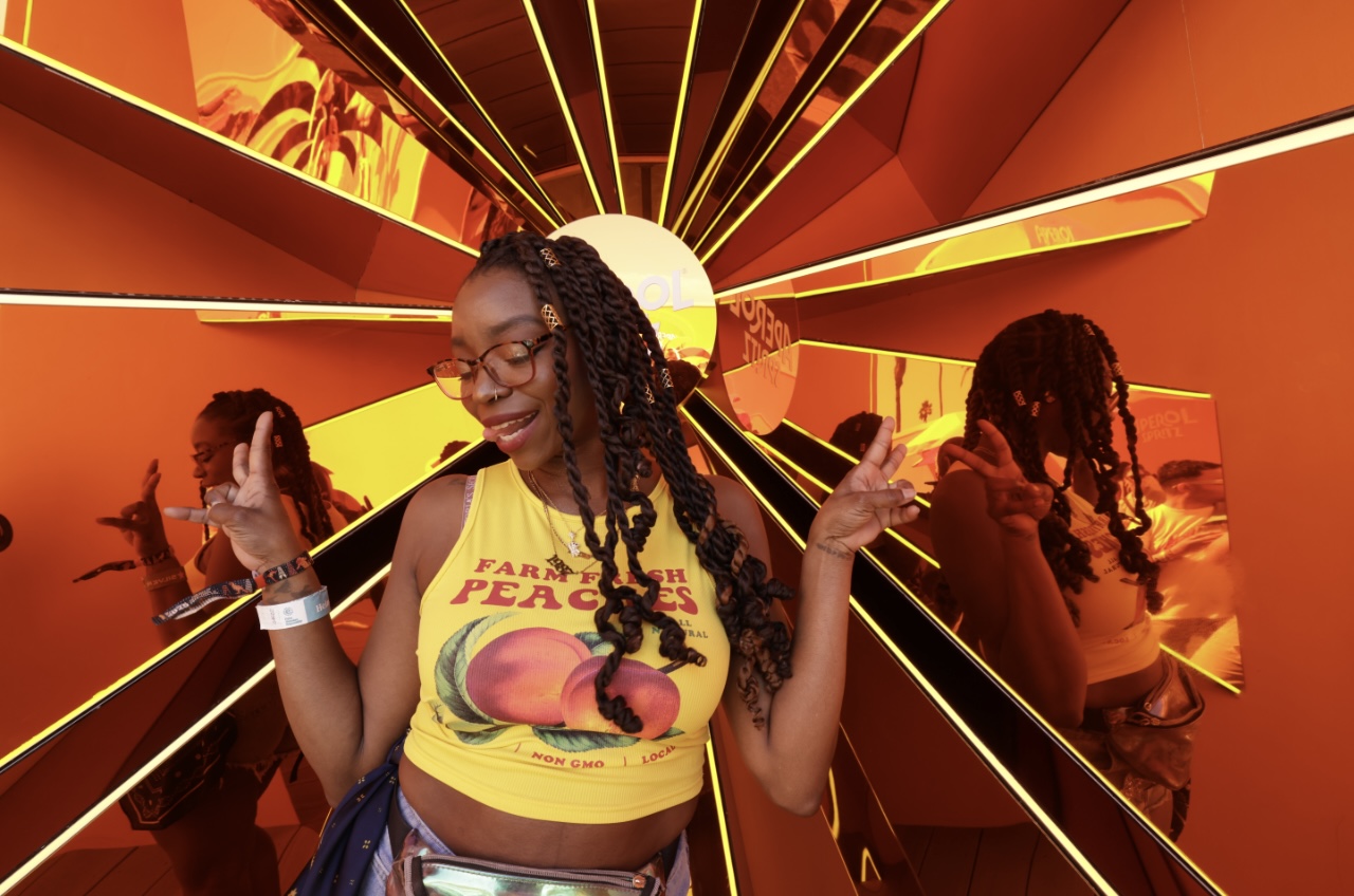 Young woman with glasses and braided hair smiling with arms raised in front of colorful, abstract background.