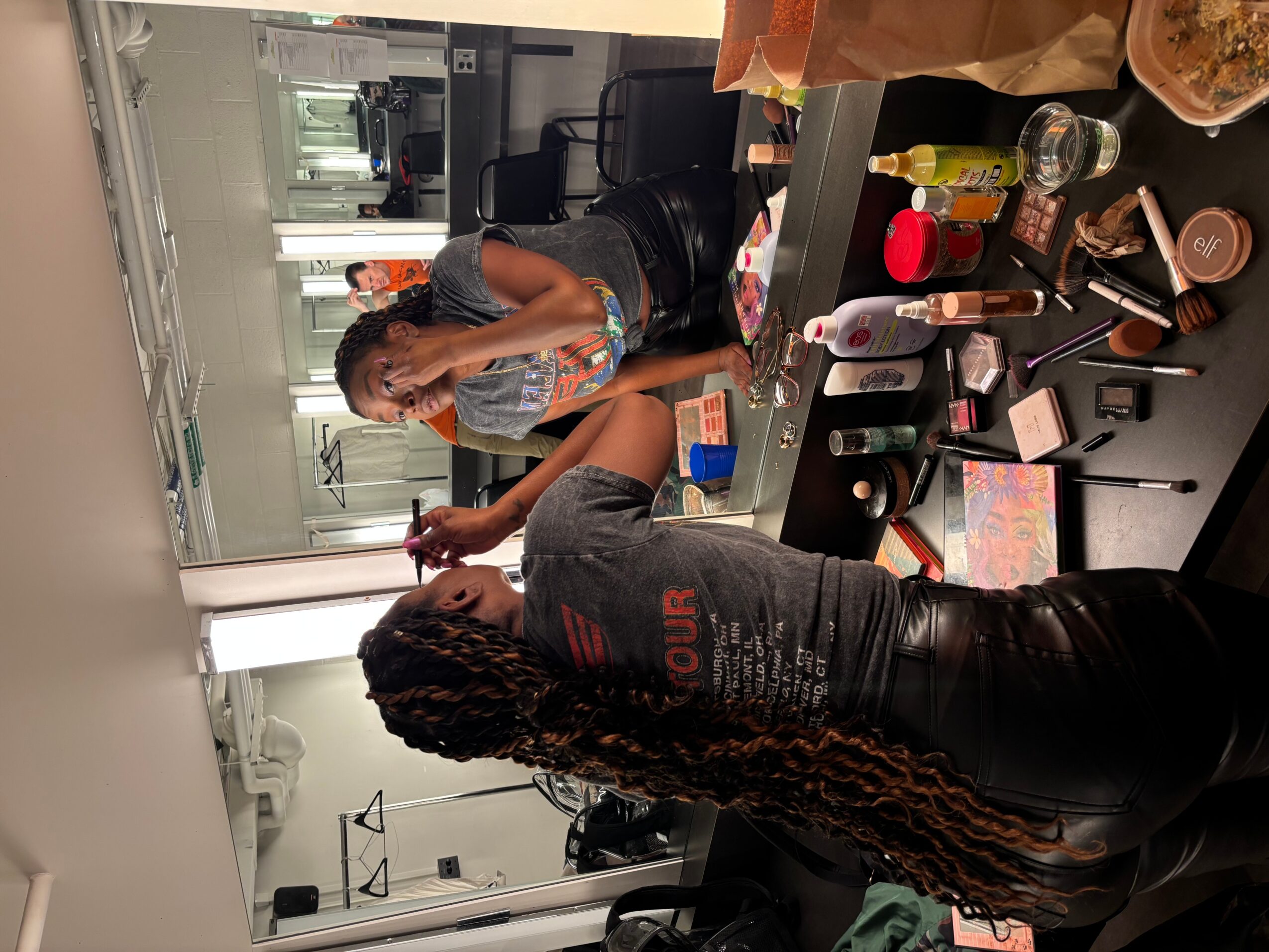 Two women in a makeup or dressing room, one sitting and the other standing, surrounded by makeup and beauty products.