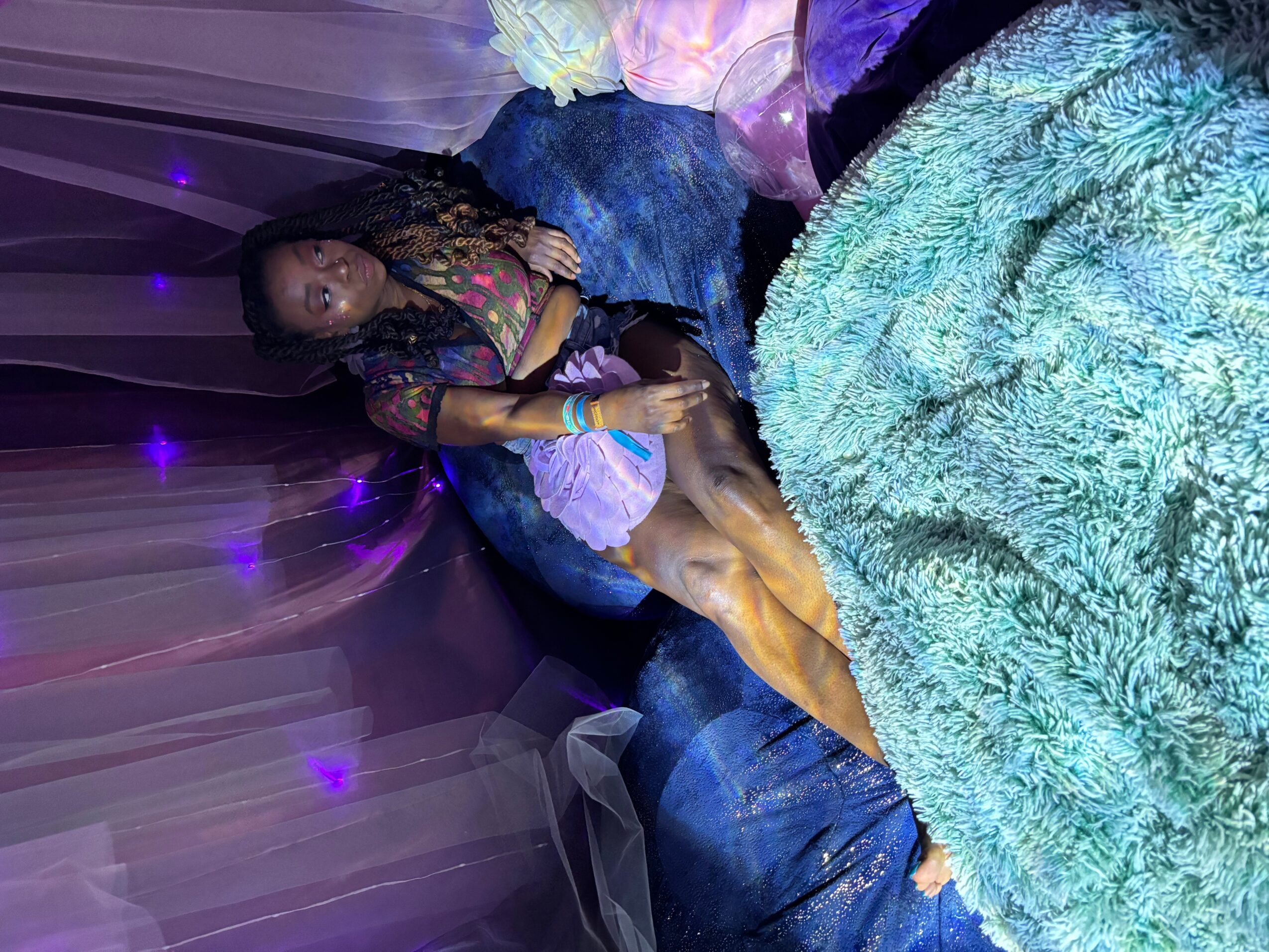 Person sitting on a bed with purple lighting, wearing a dark top and denim shorts, with a fluffy blanket nearby.
