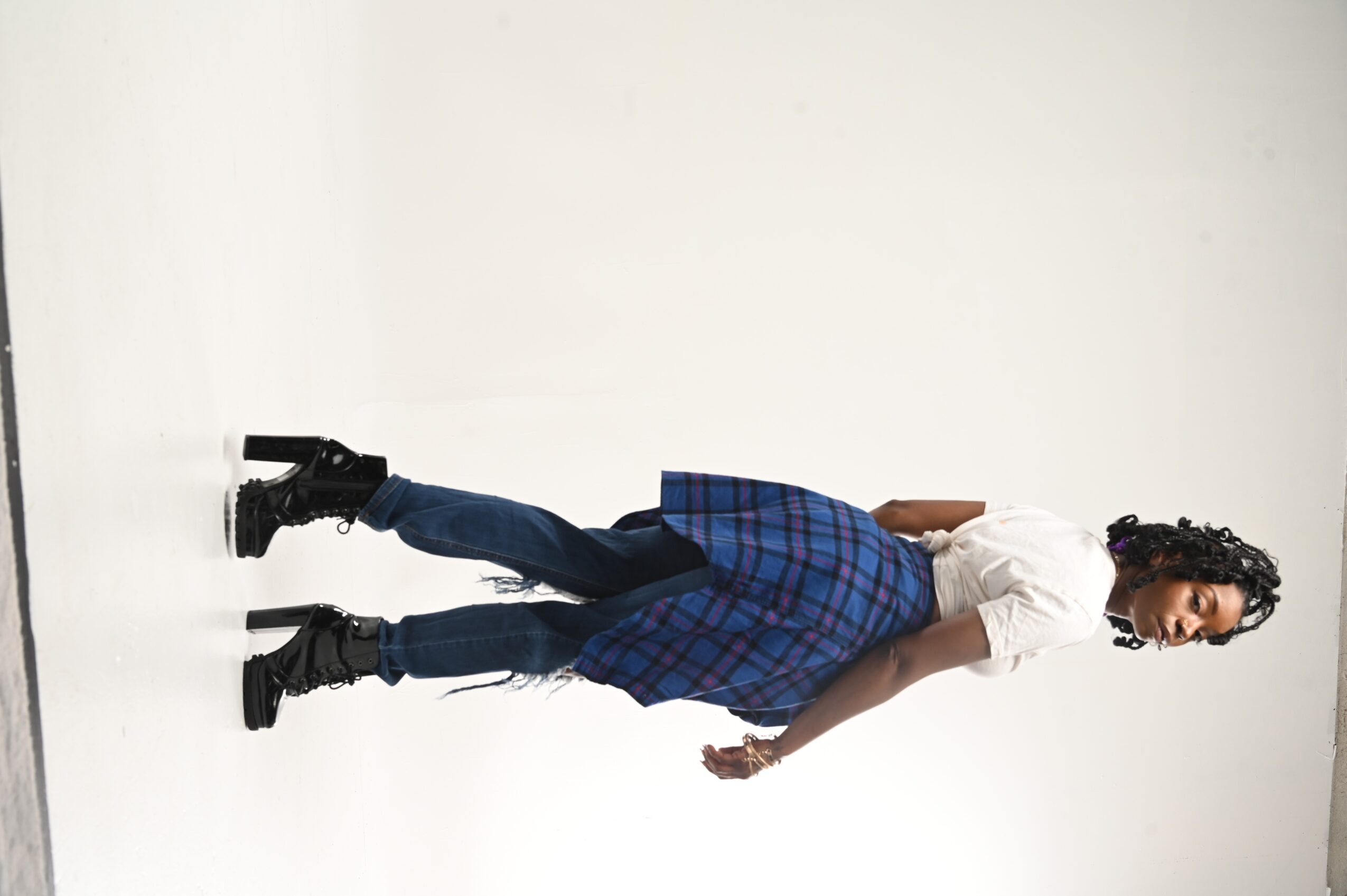 Person standing sideways against a plain white background, wearing a blue plaid dress, white blouse, and black boots.