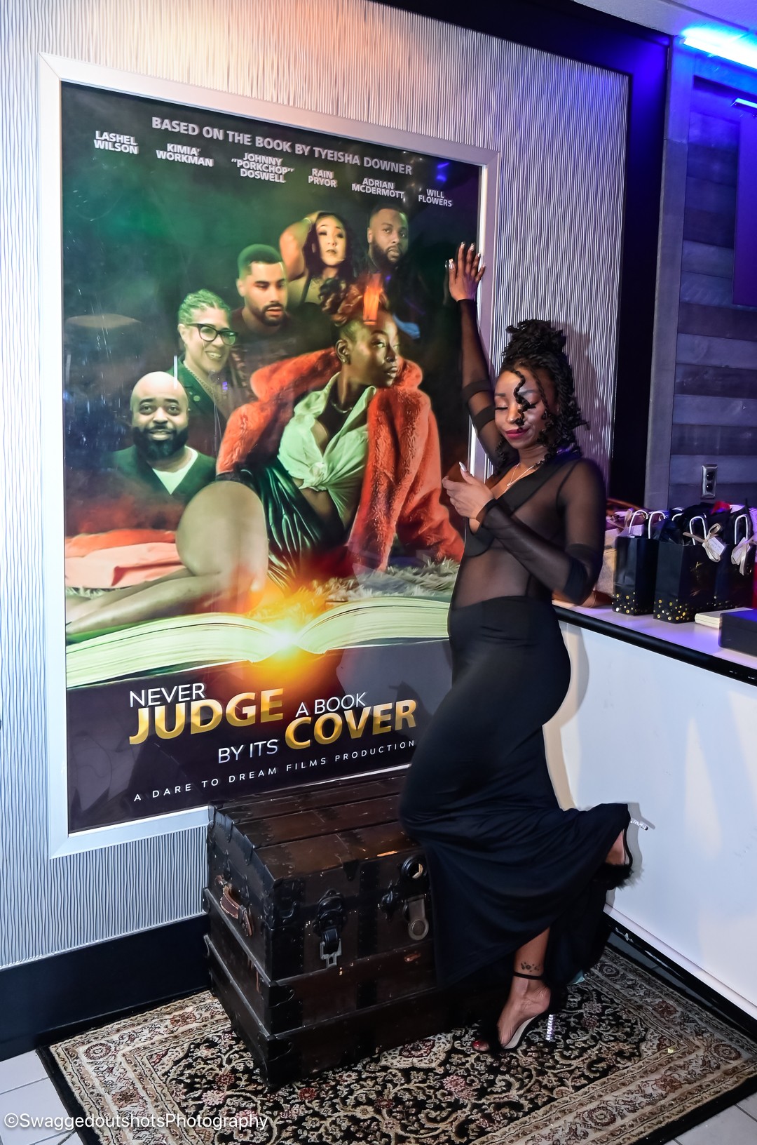 Woman in black dress standing next to a poster for the movie 'Never Judge a Book by Its Cover' in a room with a patterned rug.