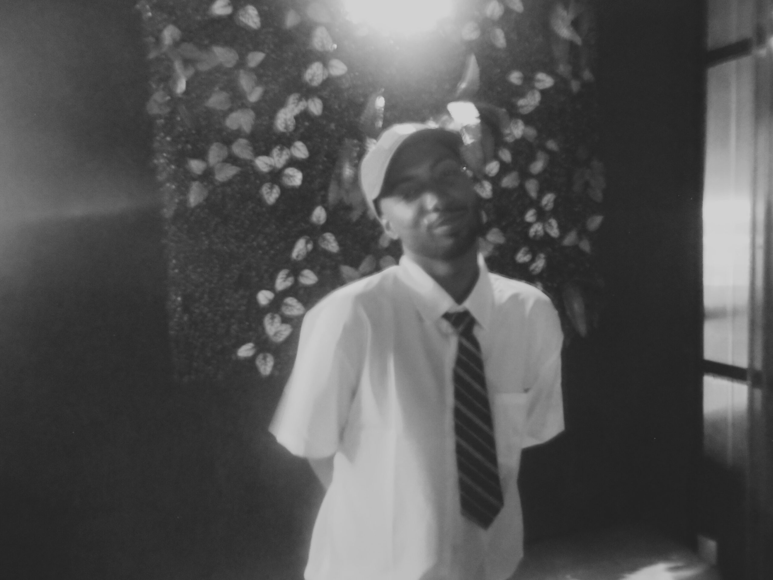 A person in a school uniform with a tie, standing in front of a floral background, smiling slightly.