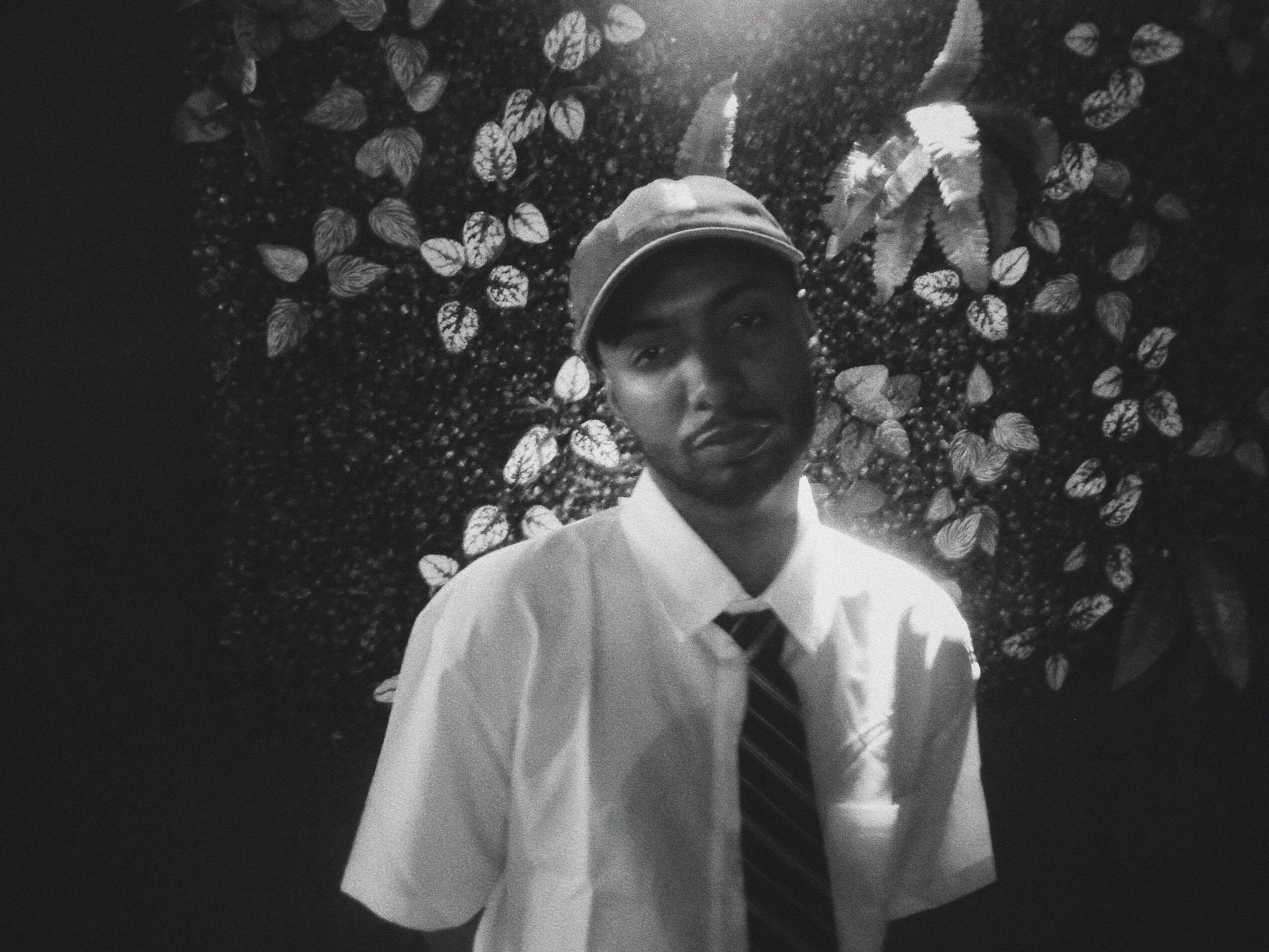 Young person wearing a cap, white shirt, and striped tie, standing in front of a leafy background.
