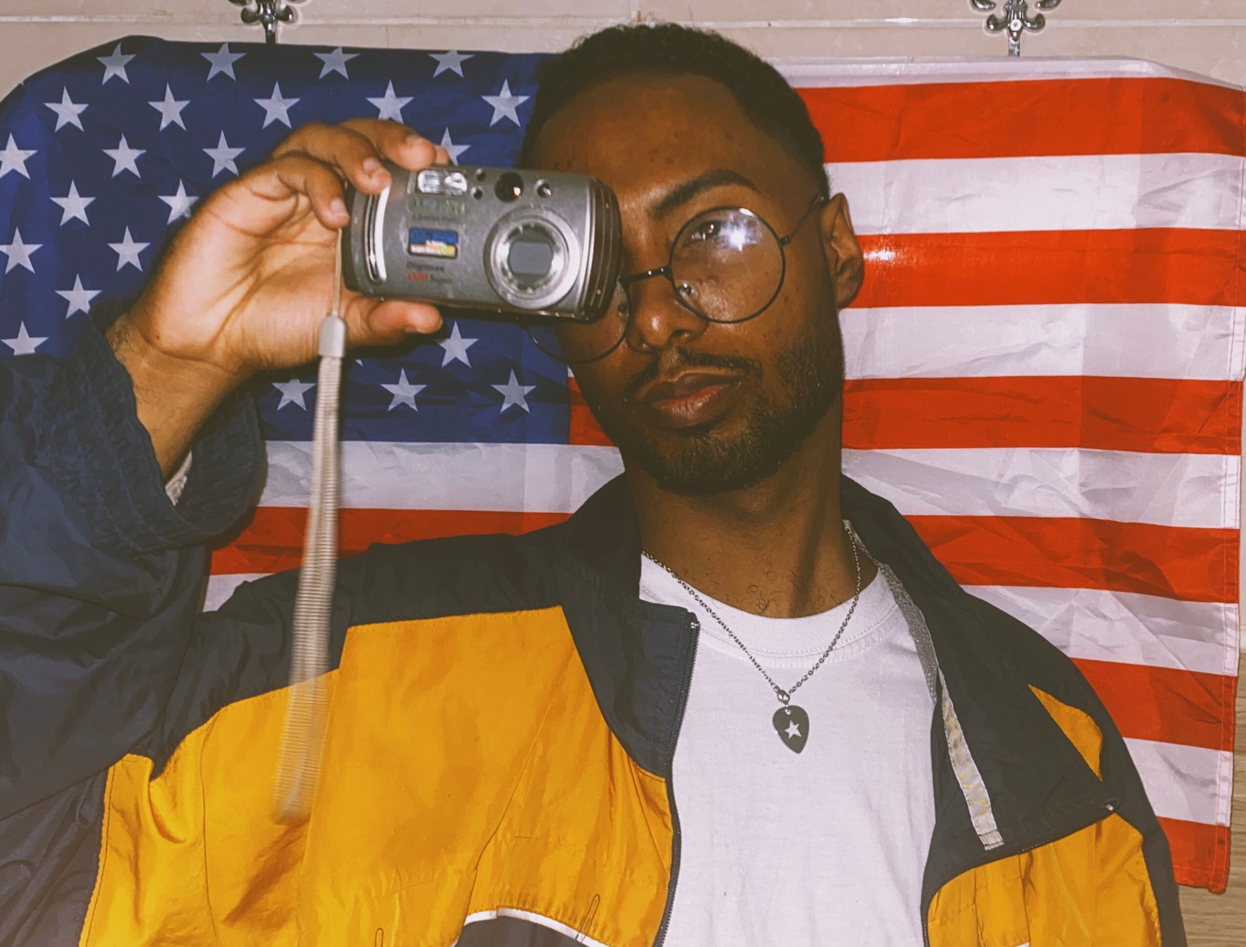 Person with glasses holding a camera in front of an American flag backdrop.