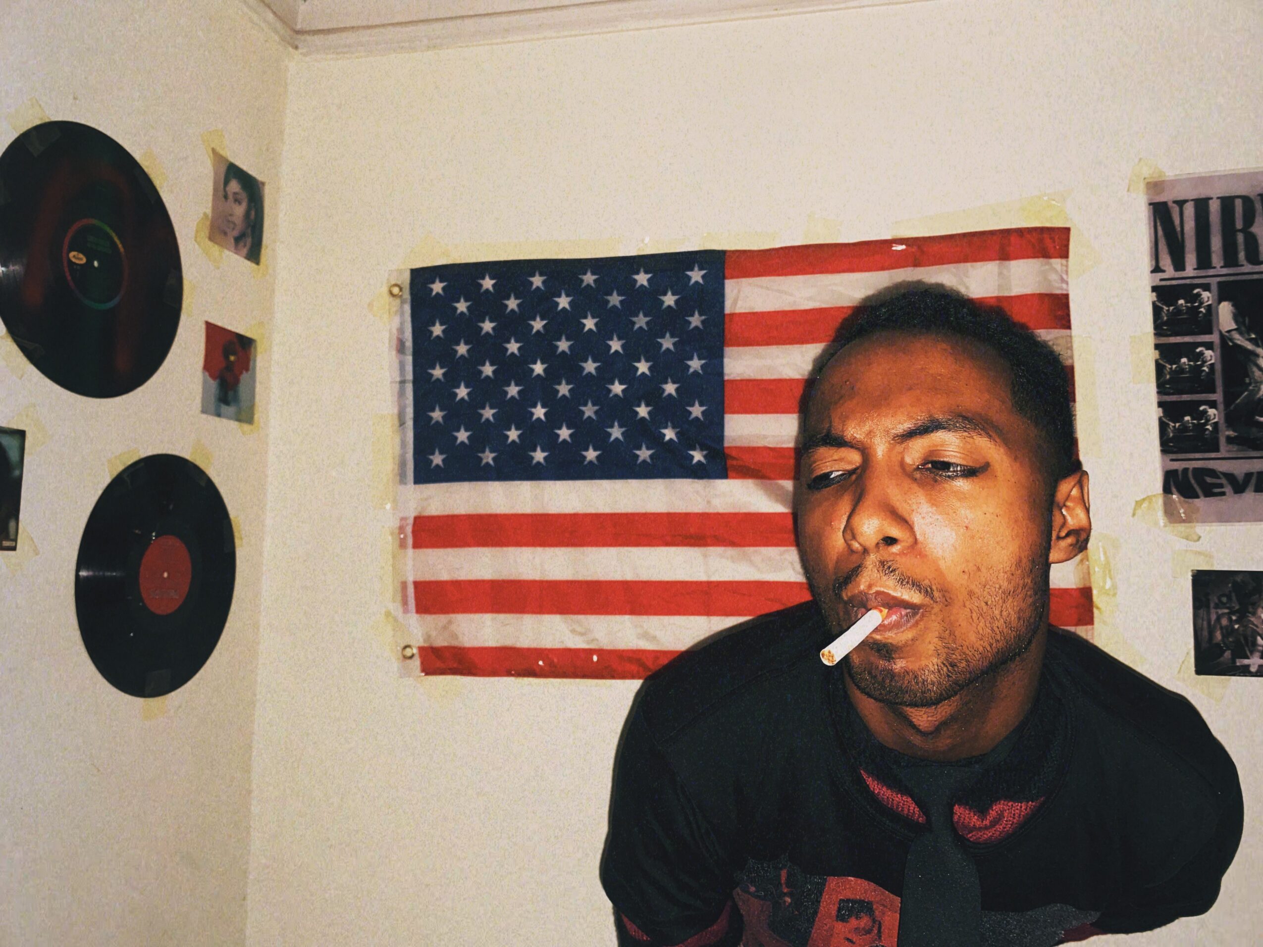 Man with a cigarette in his mouth in front of an American flag on the wall, with vinyl records and posters nearby.