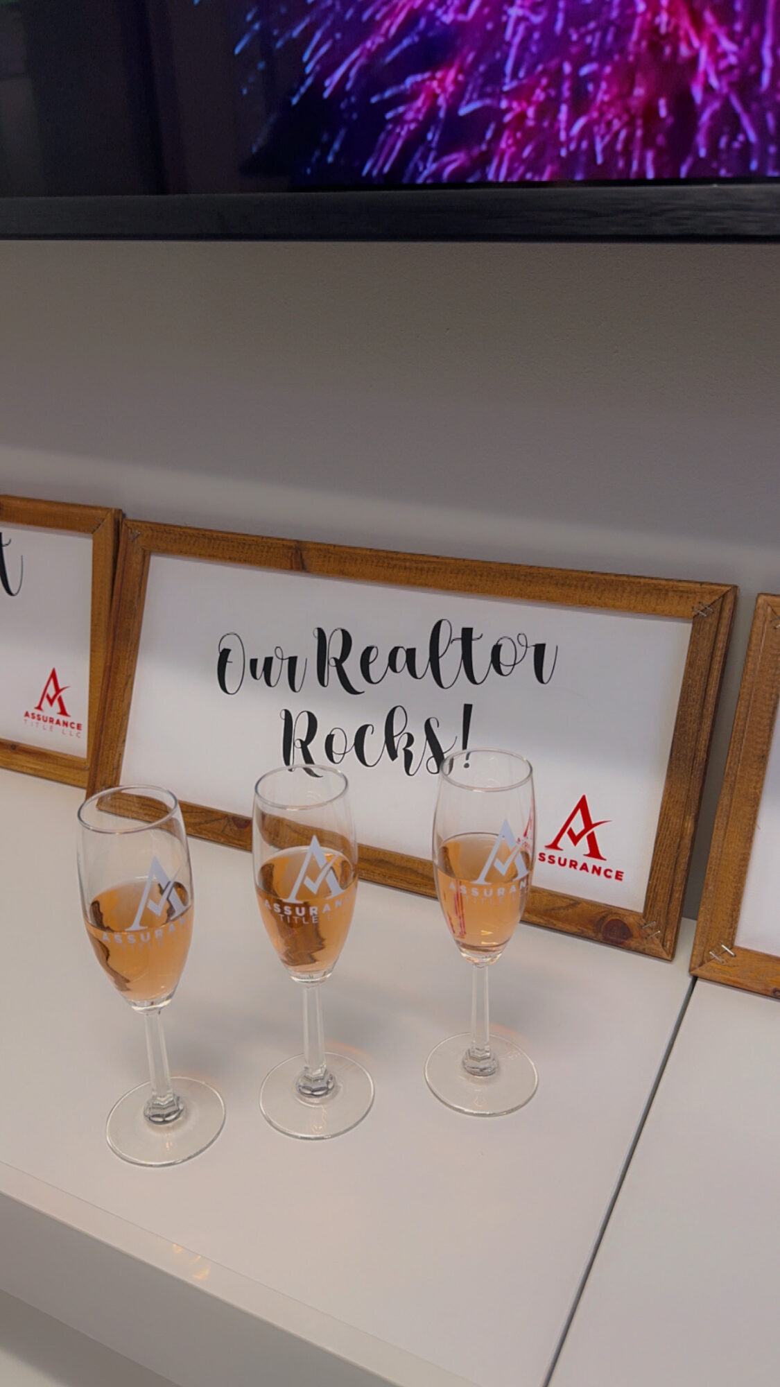 Three champagne glasses on a table in front of a framed sign that reads 'Our Realtor Rocks!'.