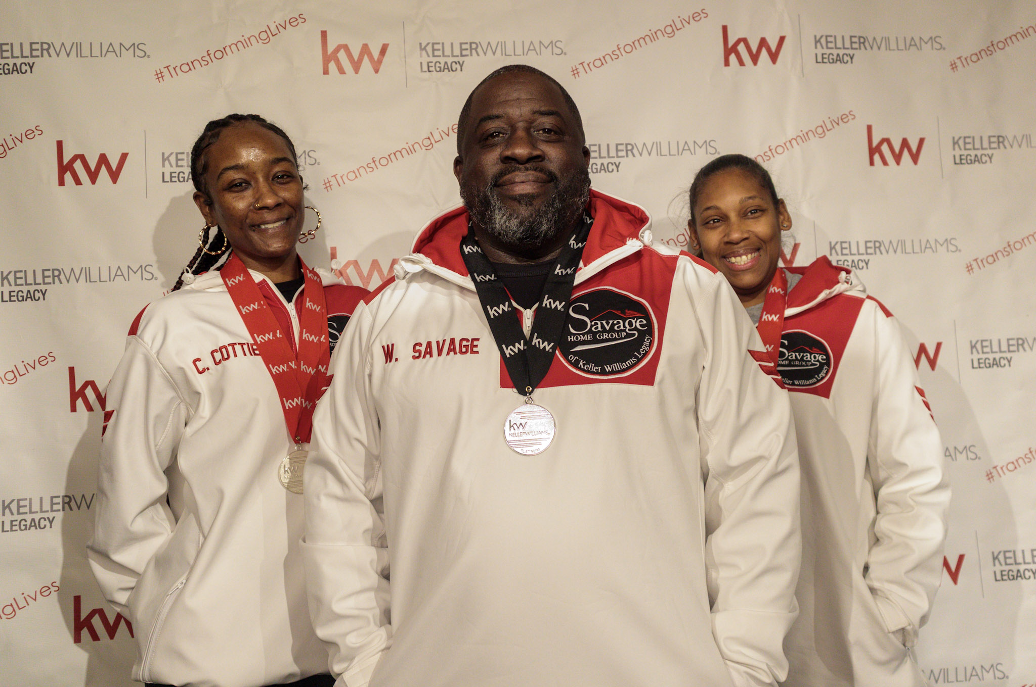 Three people wearing white jackets with red accents, standing in front of a backdrop with logos, smiling.