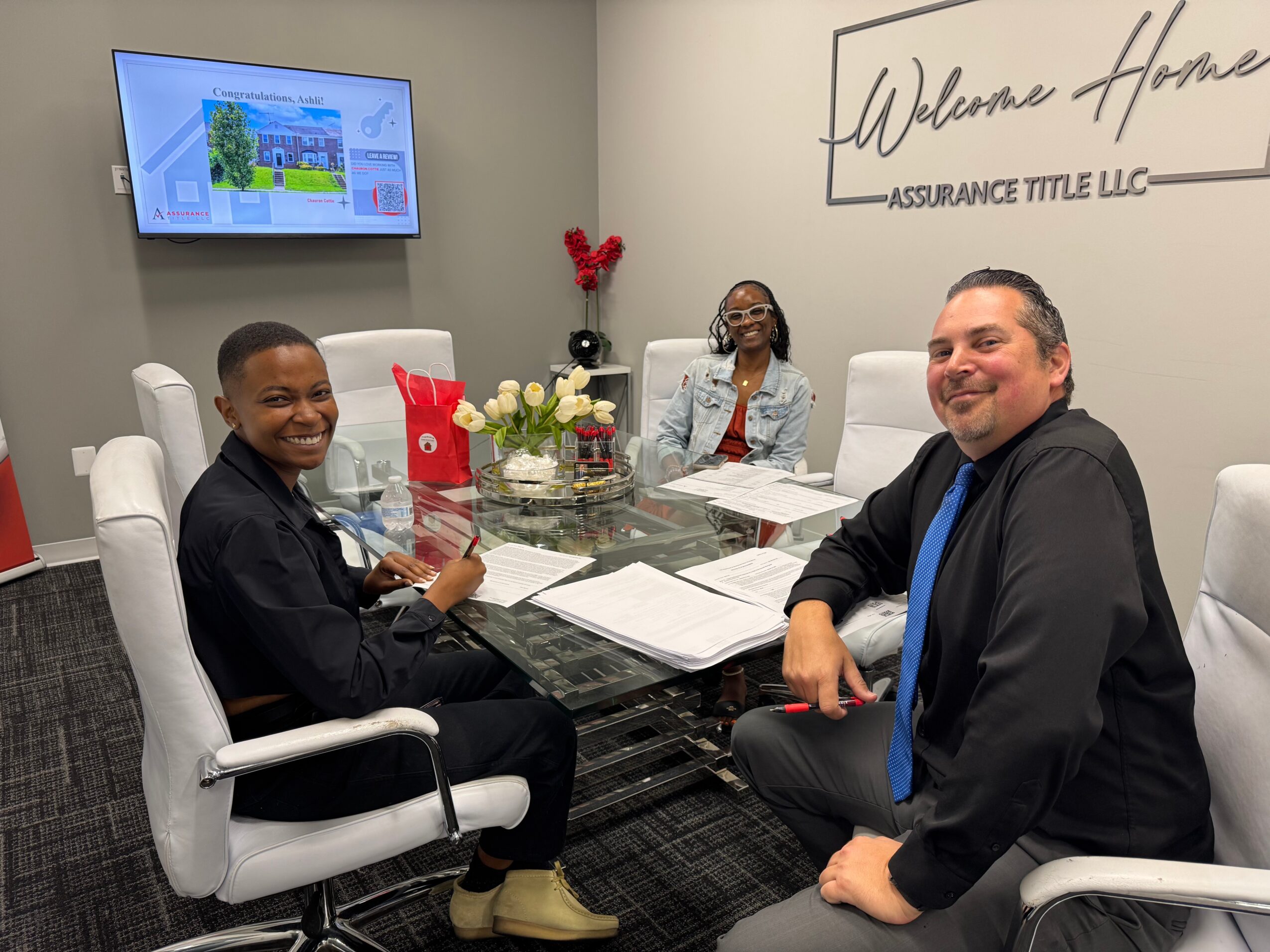 Three people sit around a conference table in a meeting room, smiling, with a TV screen and wall sign in background.