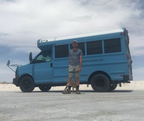 Person standing in front of a blue bus on a flat, open landscape with cloudy sky.