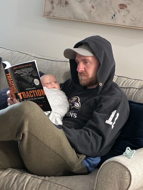 Man with a beard wearing a black hoodie and cap, sitting on a sofa, reading a book to a baby wrapped in a blanket.