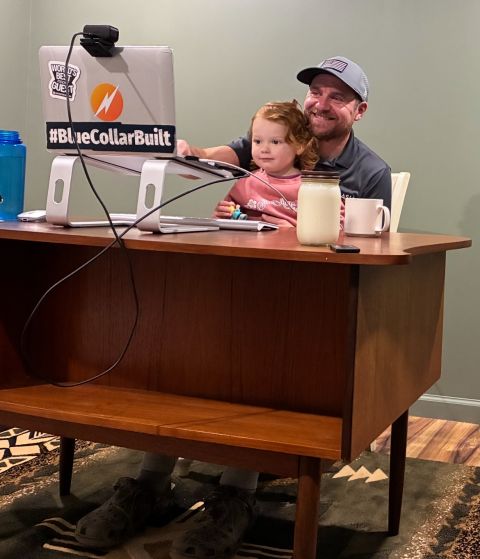 A man and a young girl sit at a wooden table with a laptop, drinks, and a sign, smiling at the camera.