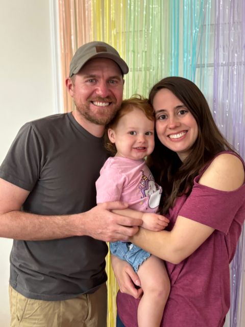 Three people, a man, a woman, and a young girl, smiling and posing together in front of colorful curtains.