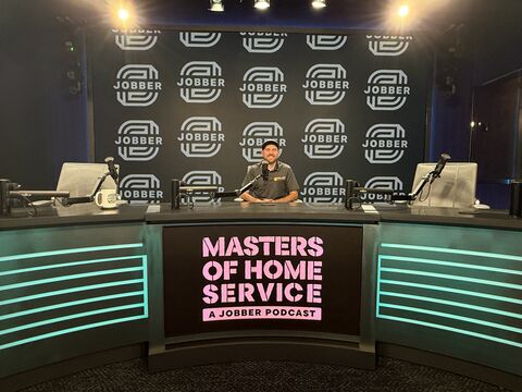 Person sitting at a desk on a stage with a backdrop and a sign reading 'Masters of Home Service'.