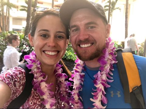 Two smiling people wearing leis, outdoors with palm trees and buildings in background.