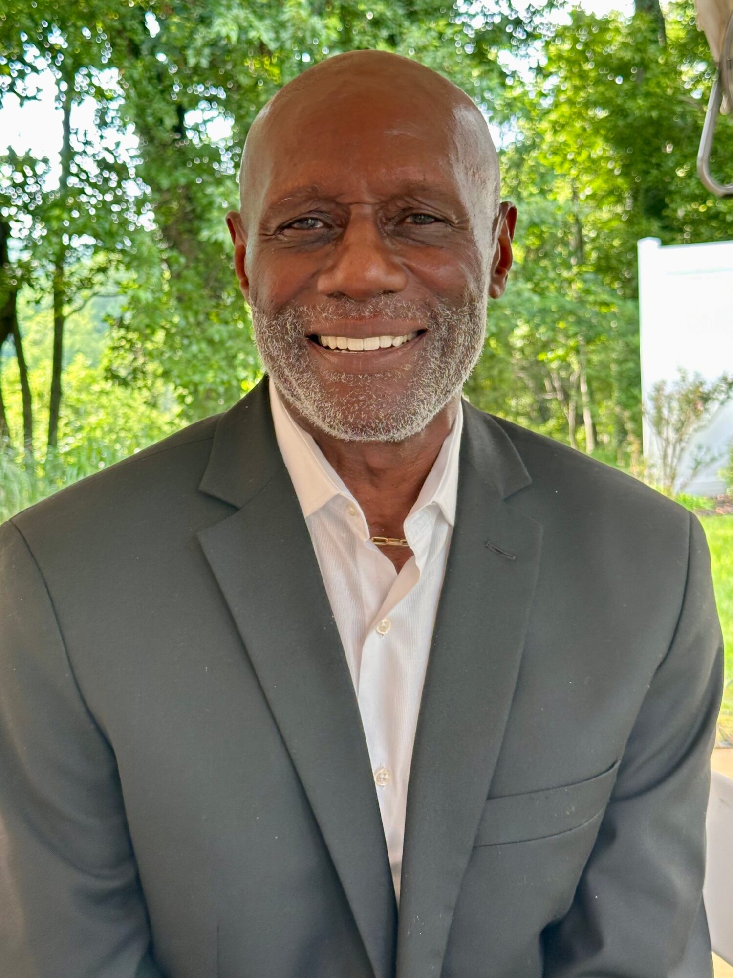 Smiling man in a suit outdoors with trees in the background.