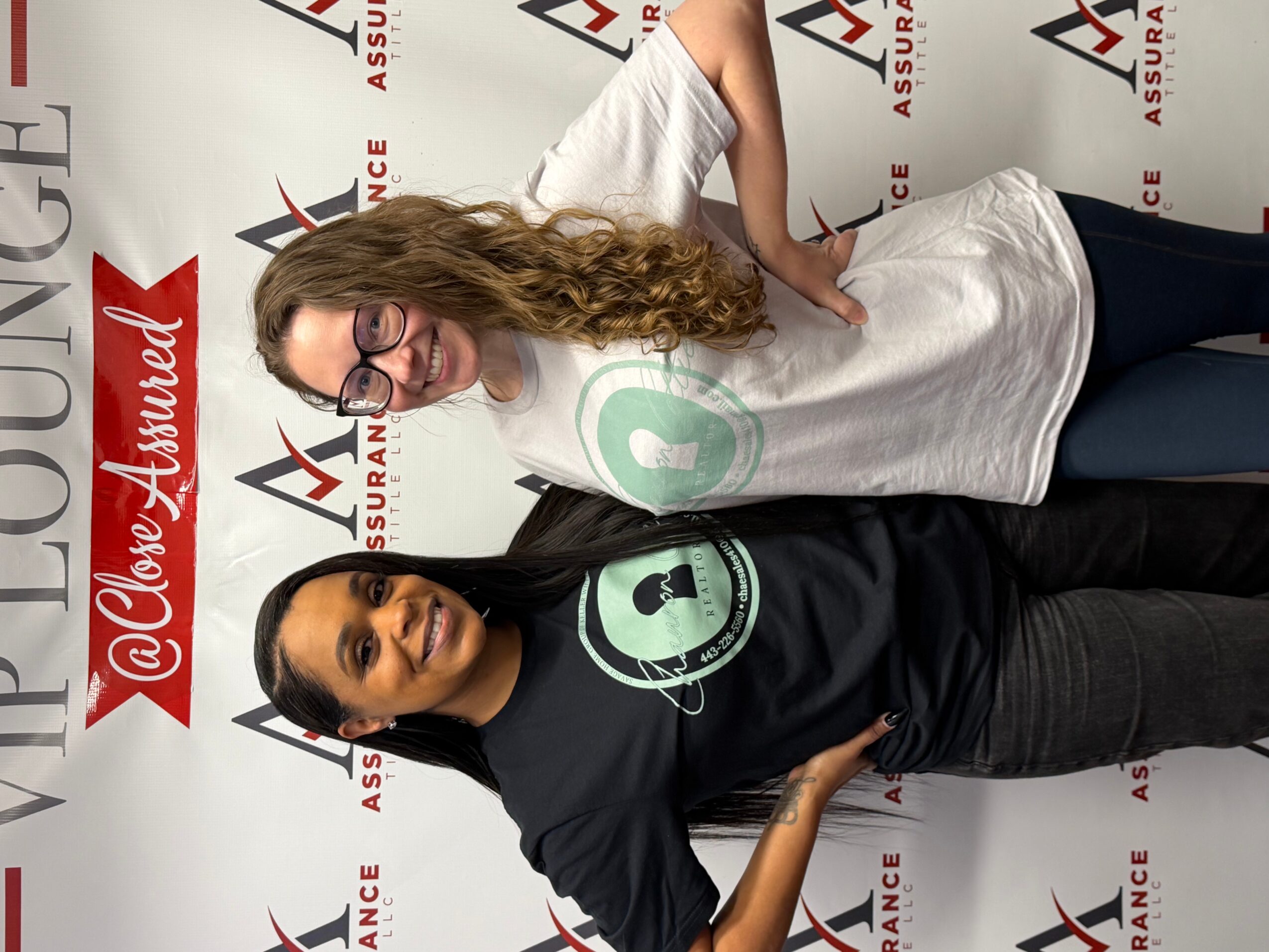 Two women standing side by side in front of a backdrop with logos, smiling and posing for the camera.