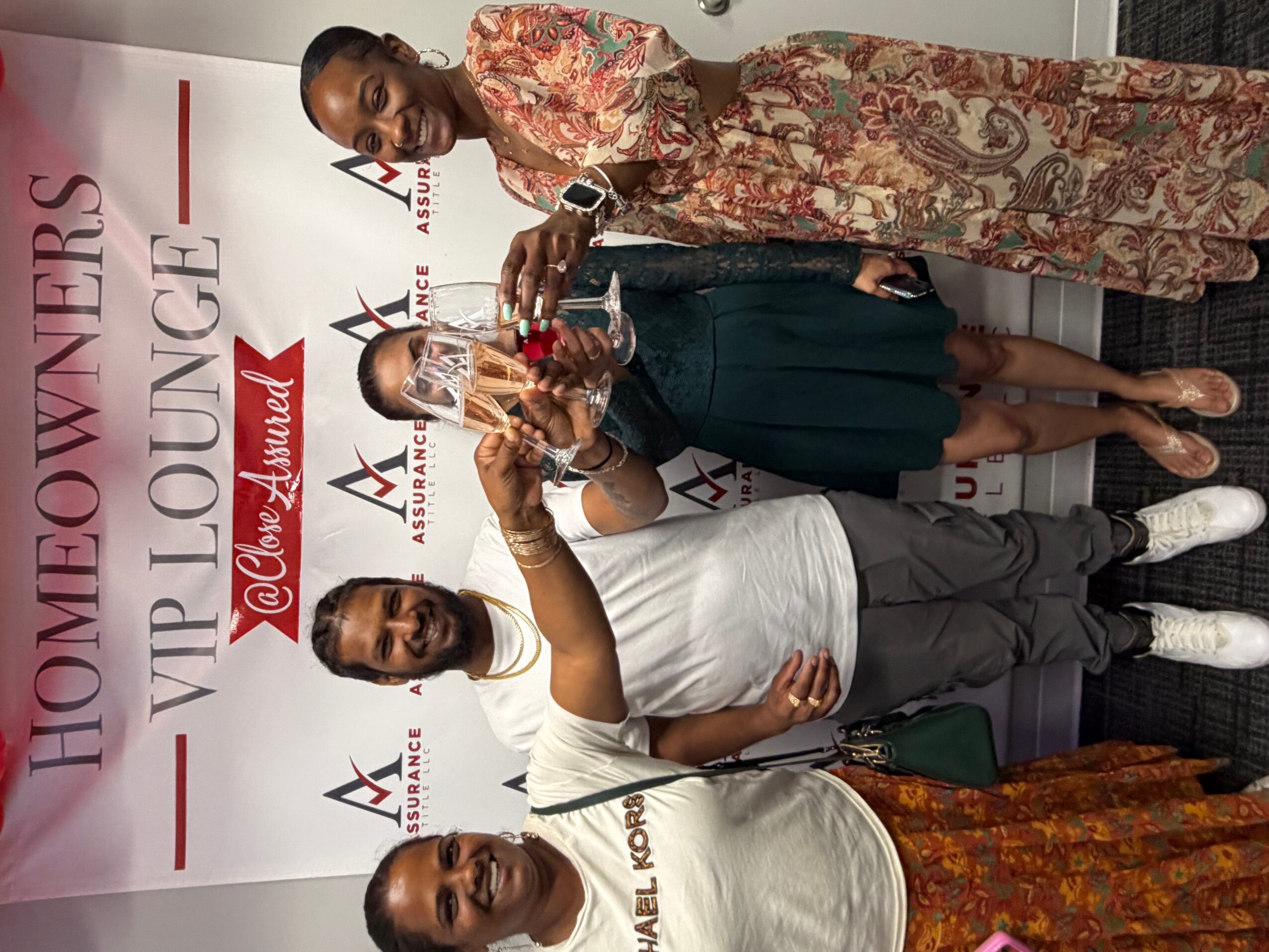 Four women standing together, one holding a glass, in front of a banner with event details.