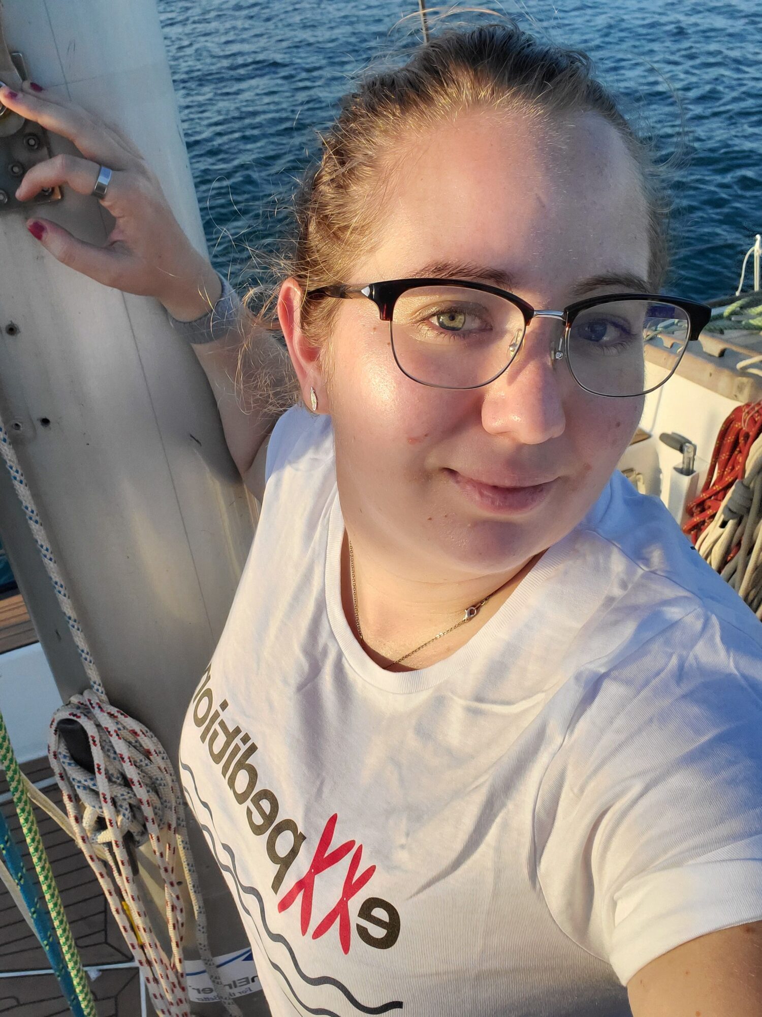 Person with glasses on a boat, water in background, wearing a white shirt, smiling, holding a rope.