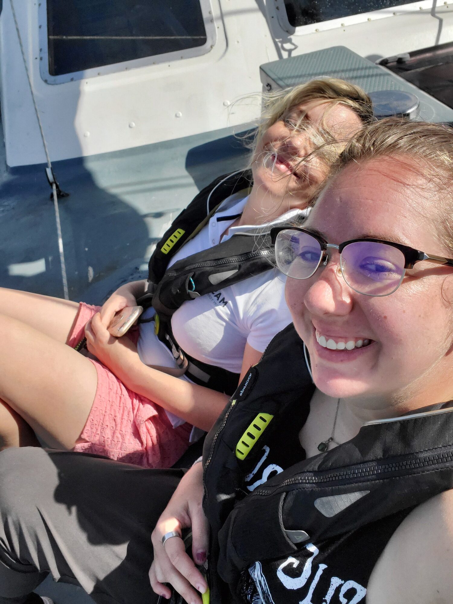 Two smiling women wearing glasses and harnesses, sitting outdoors on a sunny day.