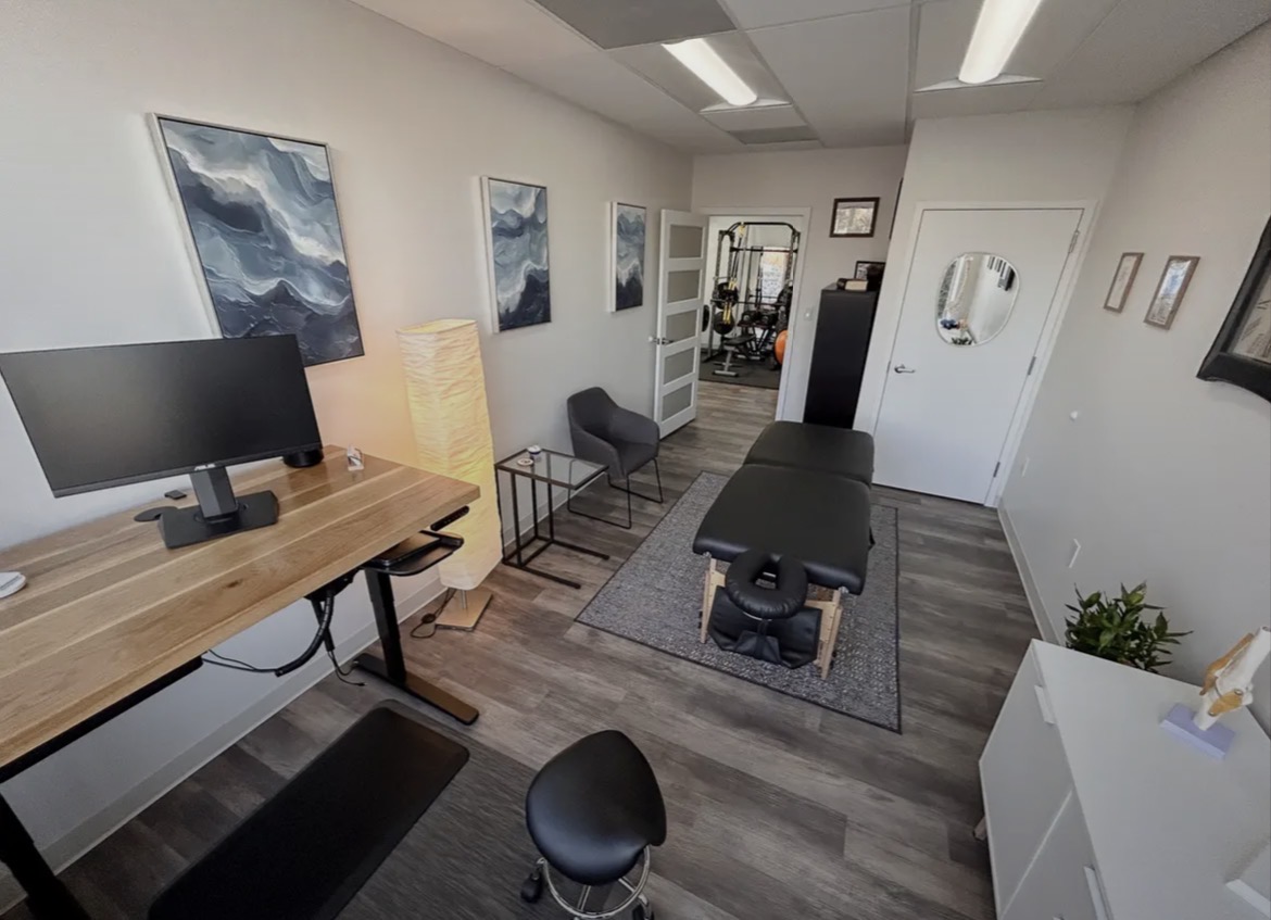 Massage table in a room with artwork, a desk with a monitor, and a chair, set up for therapy or treatment.