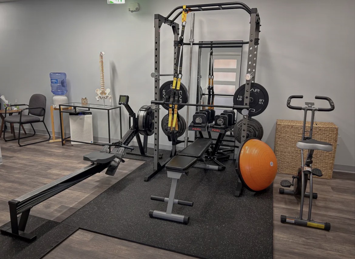 Gym equipment including a squat rack, weight plates, a bench, a stability ball, and a rowing machine in a workout room.