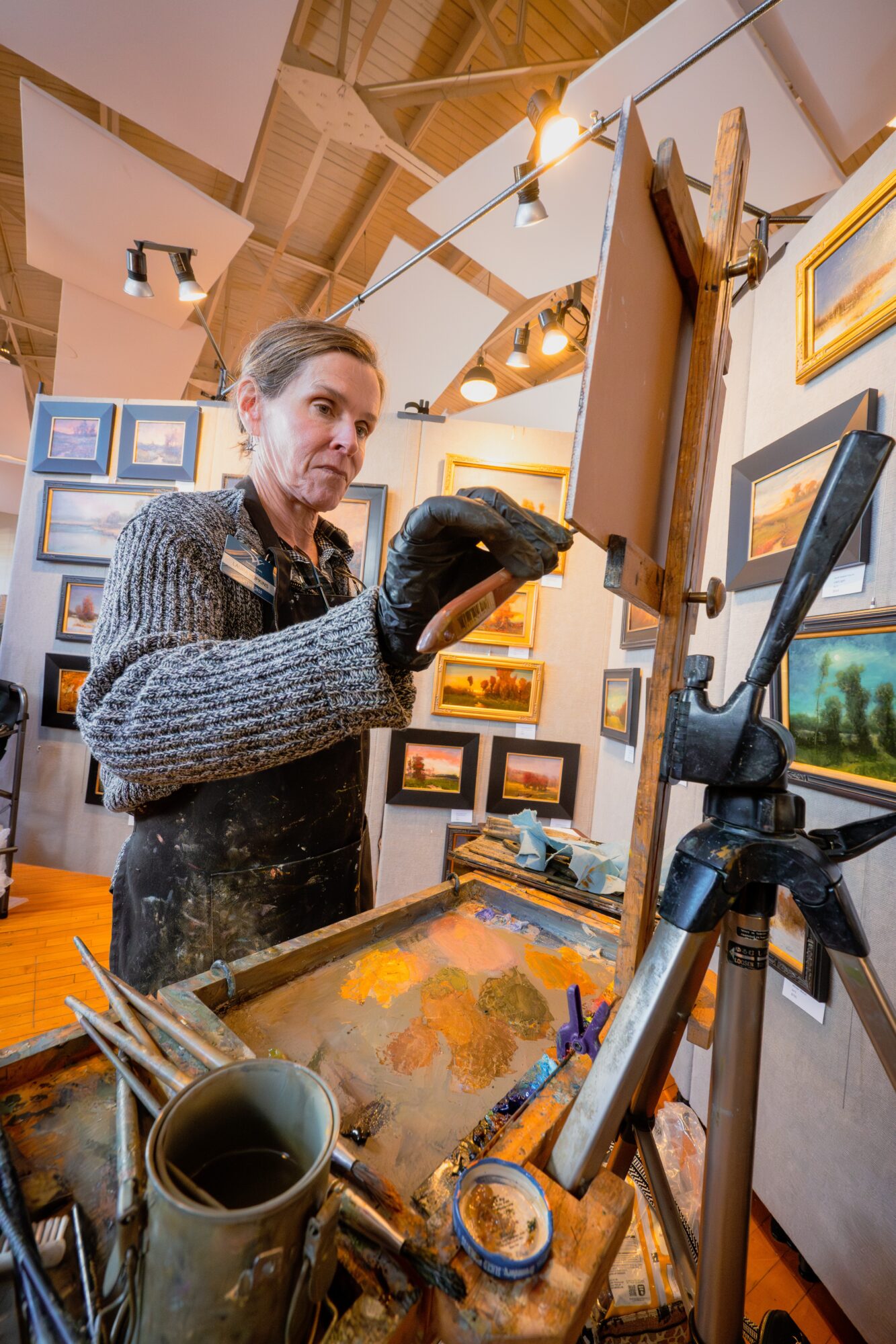 Woman painting on canvas in art gallery, surrounded by framed artwork, with painting supplies on table.