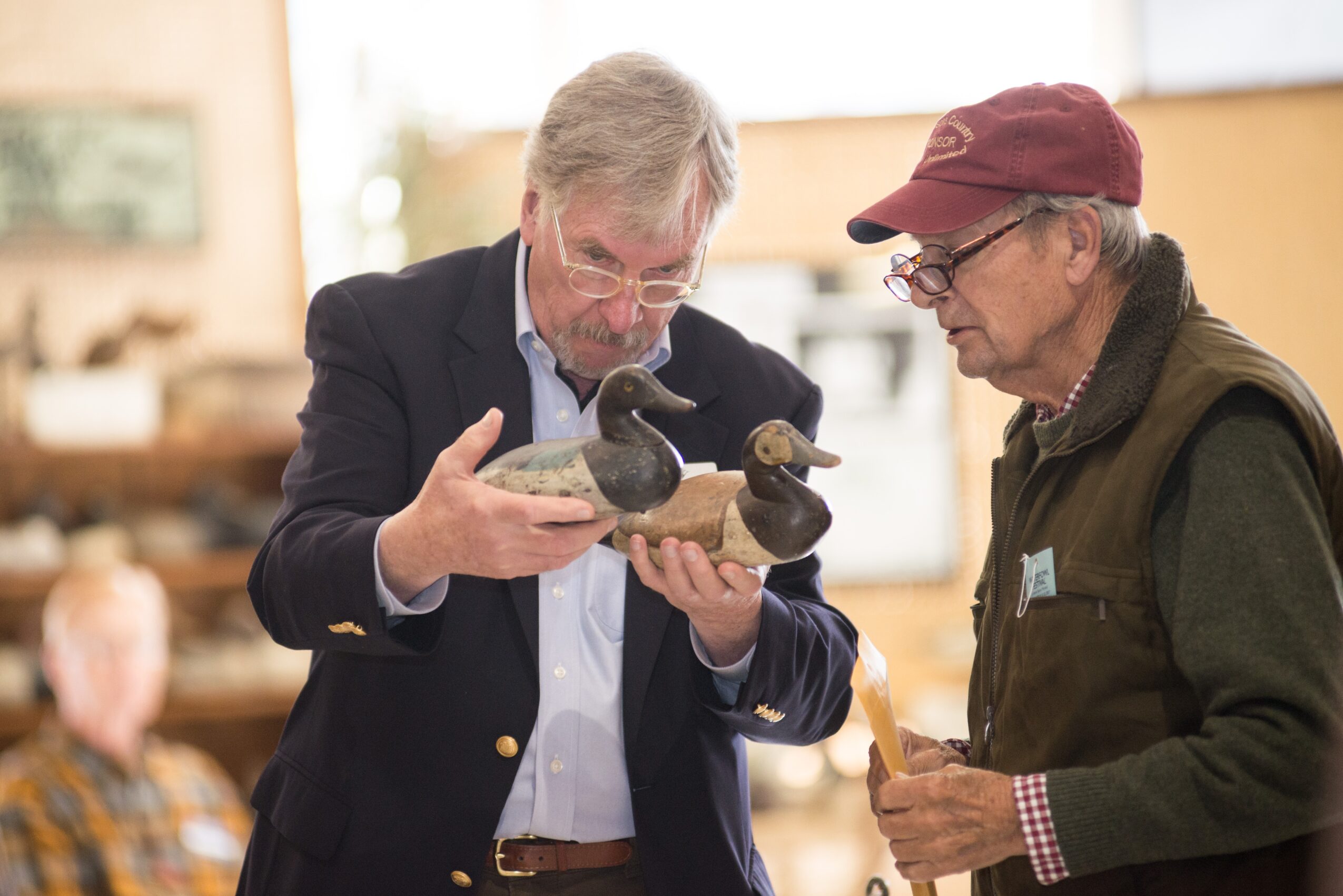 Two men examine a small sculpture or artifact in a well-lit indoor setting, with blurred background and other people present.