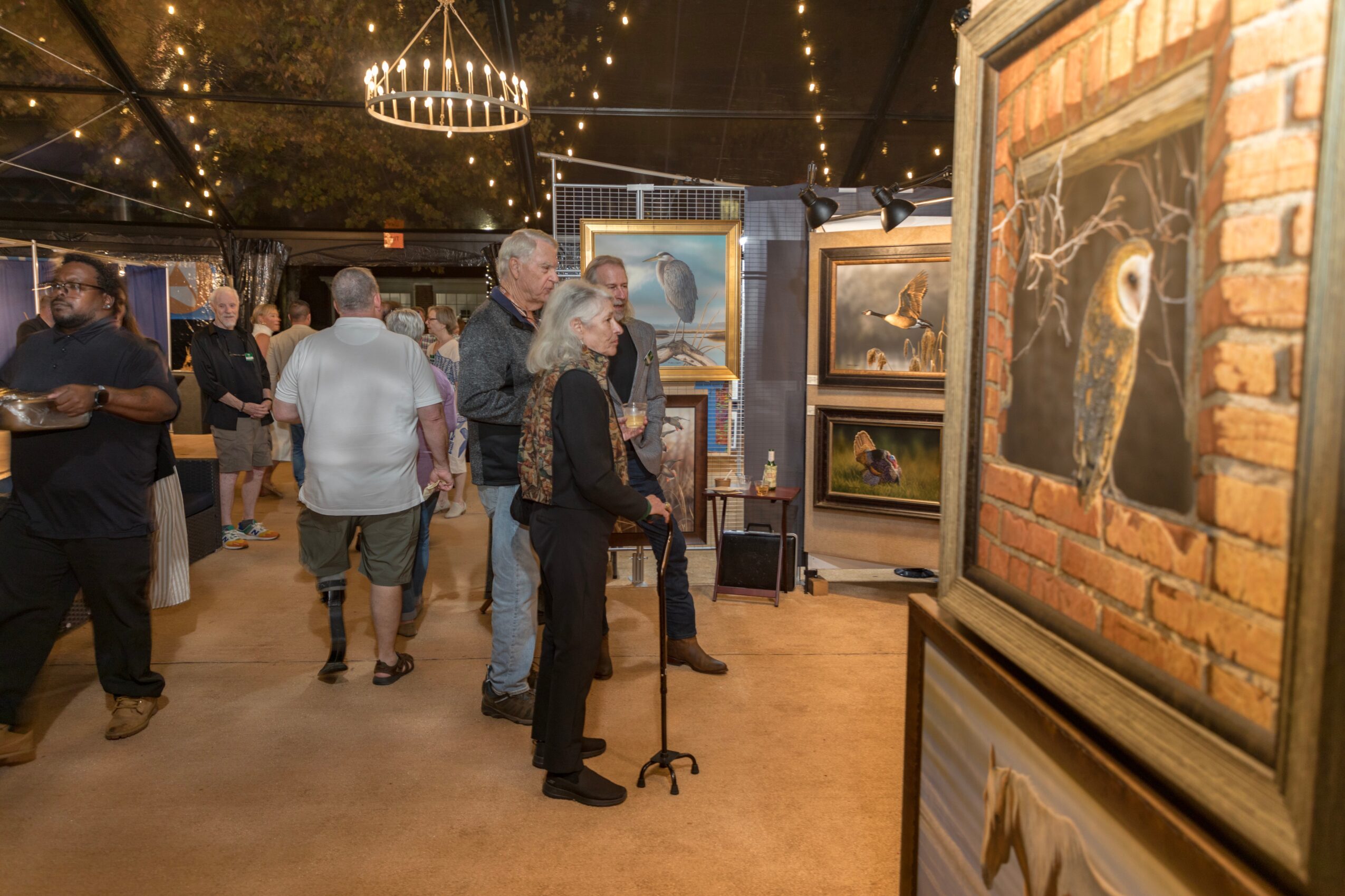 People viewing framed paintings at an art exhibition in a well-lit indoor space with brick walls and string lights.