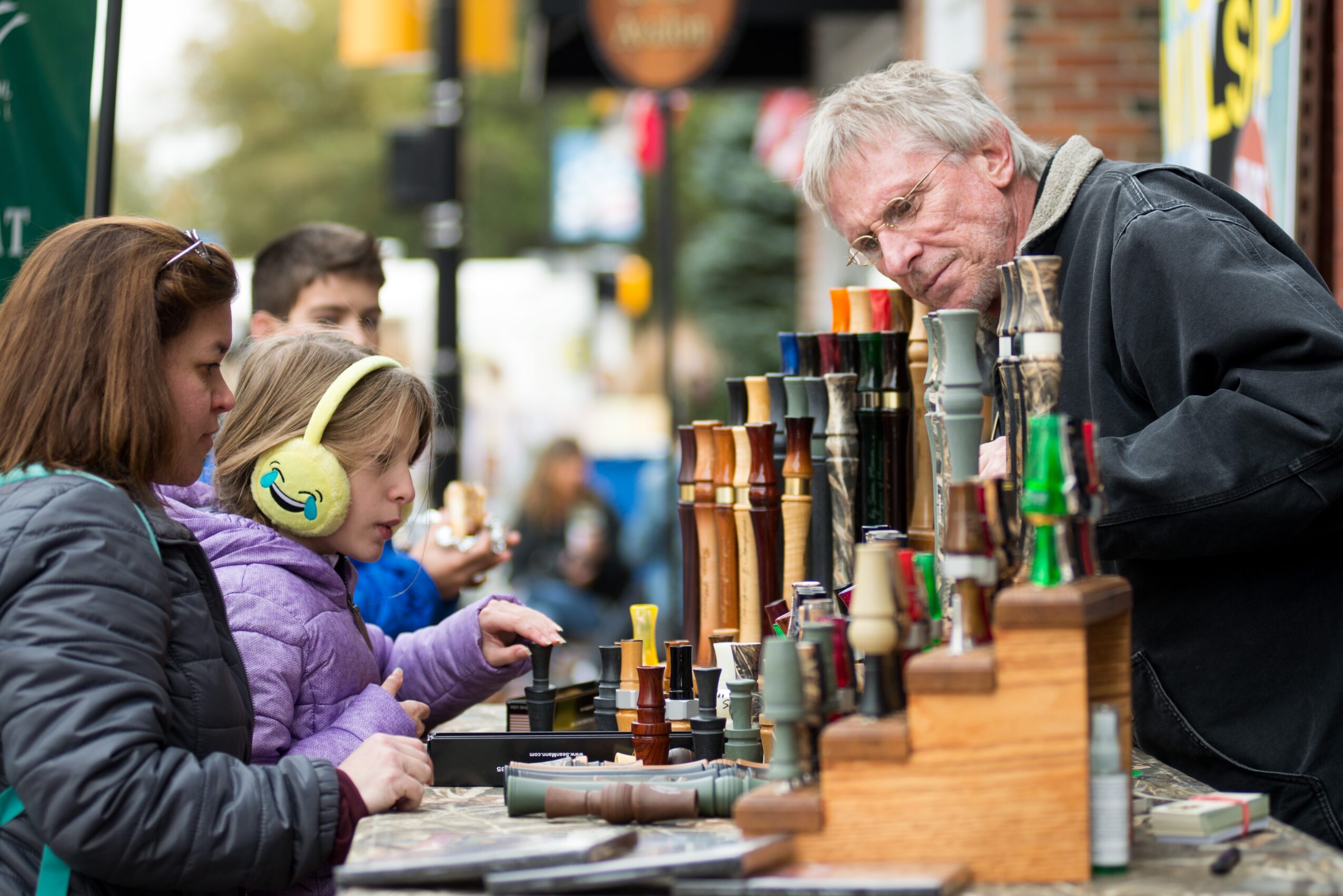 Older man and two young girls browsing colorful stacked cups at an outdoor market stall.