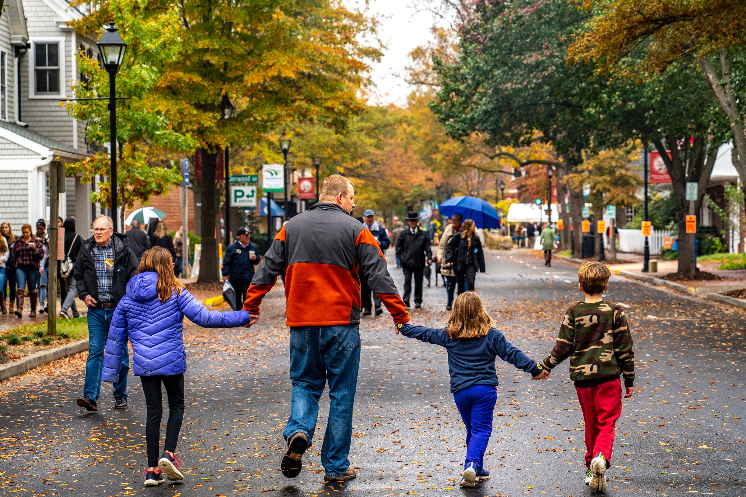 People walk on a tree-lined street holding hands, with colorful autumn leaves and buildings in the background.